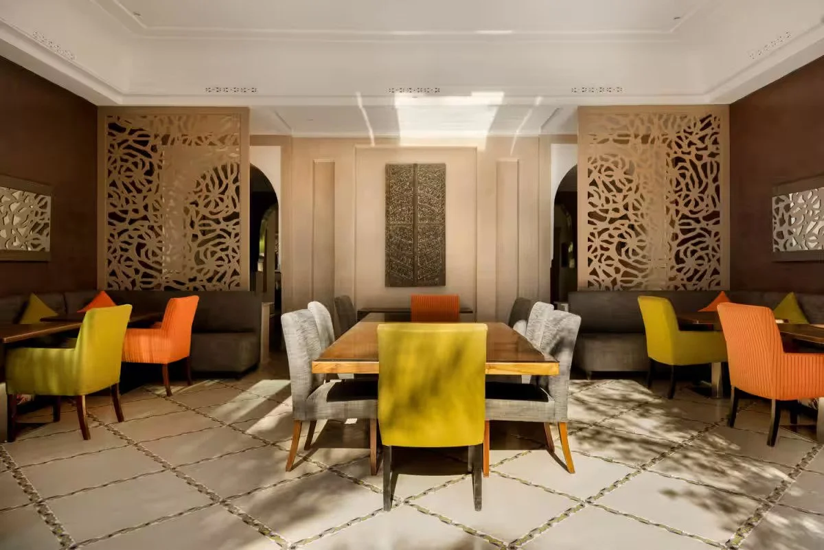 Salle à manger élégante avec une grande table en bois, chaises colorées, murs en taupe et panneaux décoratifs en bois sculpté. Mur central avec une œuvre d'art ornementale. Retraite pour femmes a marrakech au maroc