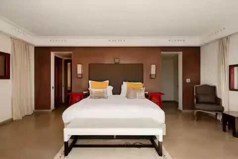 Chambre d'hôtel moderne avec grand lit, murs marrons, deux lampes murales, table de nuit rouges, fauteuil gris, fenêtres avec rideaux blancs.Retraite pour femmes a marrakech au maroc
