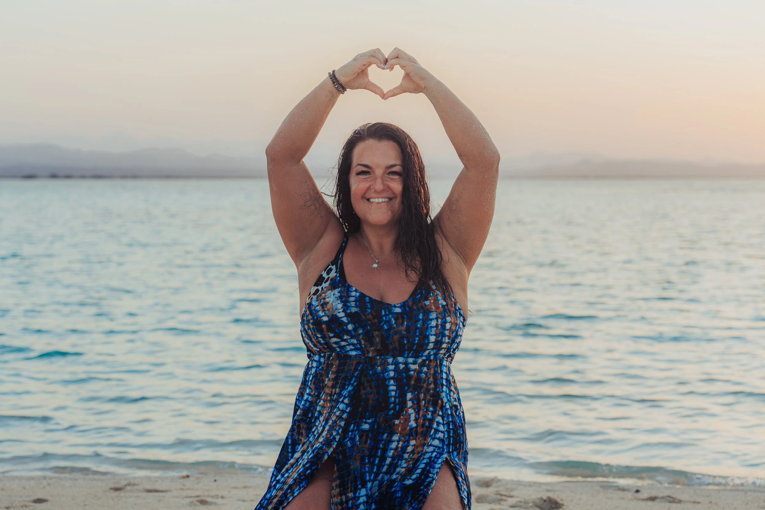 A woman at the beach making a heart shape with her hands above her head, facing the camera, wearing a blue patterned dress, smiling, with water and distant mountains in the background during sunset.