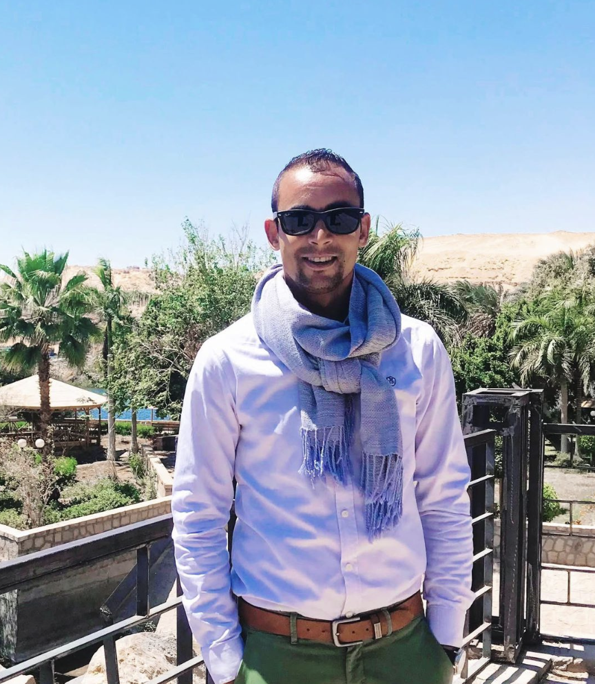 A man wearing sunglasses, a white shirt, a light blue scarf, and green pants standing outdoors with palm trees and a blue sky in the background.