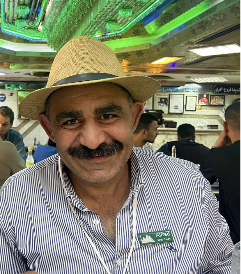 A man with a mustache wearing a straw hat and striped shirt, smiling in a crowded restaurant with green lighting and framed pictures on the wall.