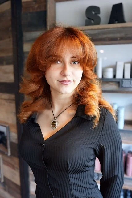 Young woman with red hair and blue eyes wearing a black striped shirt and a pendant necklace, standing indoors near wooden shelves.