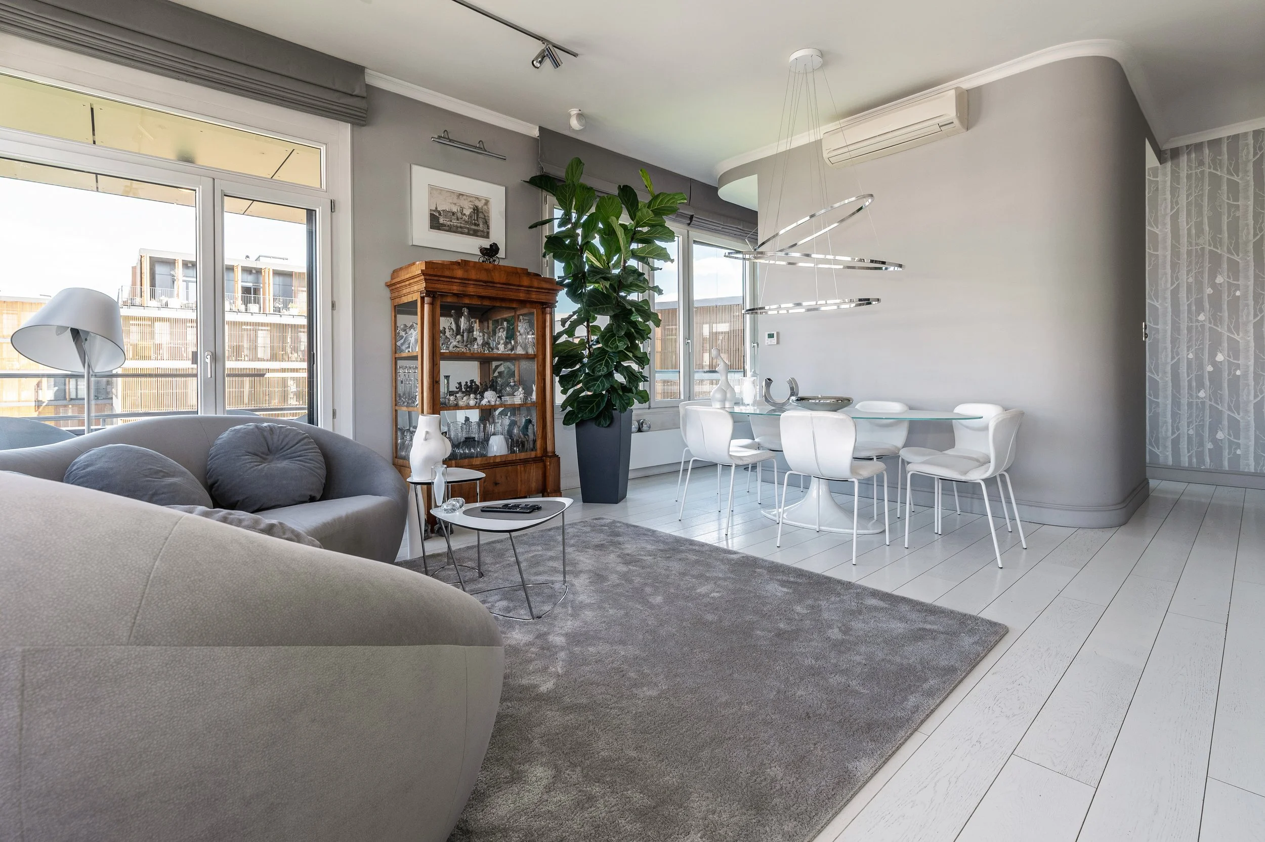 Jasny salon apartamentu w Warszawie z szarą sofą, dużymi oknami i strefą jadalnianą.