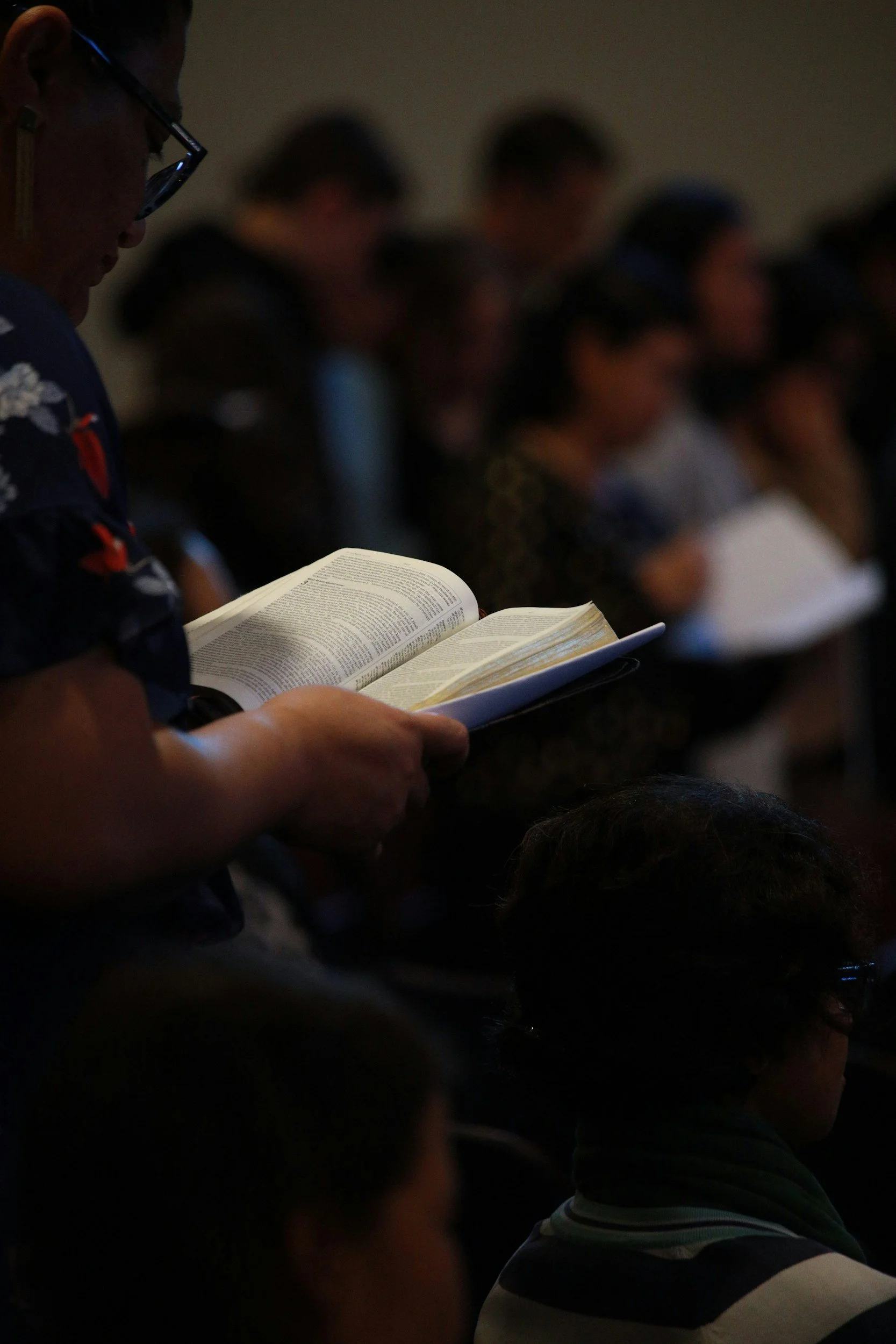 A person reading a book or a bible during a gathering or event, with other people standing or seated in the background.