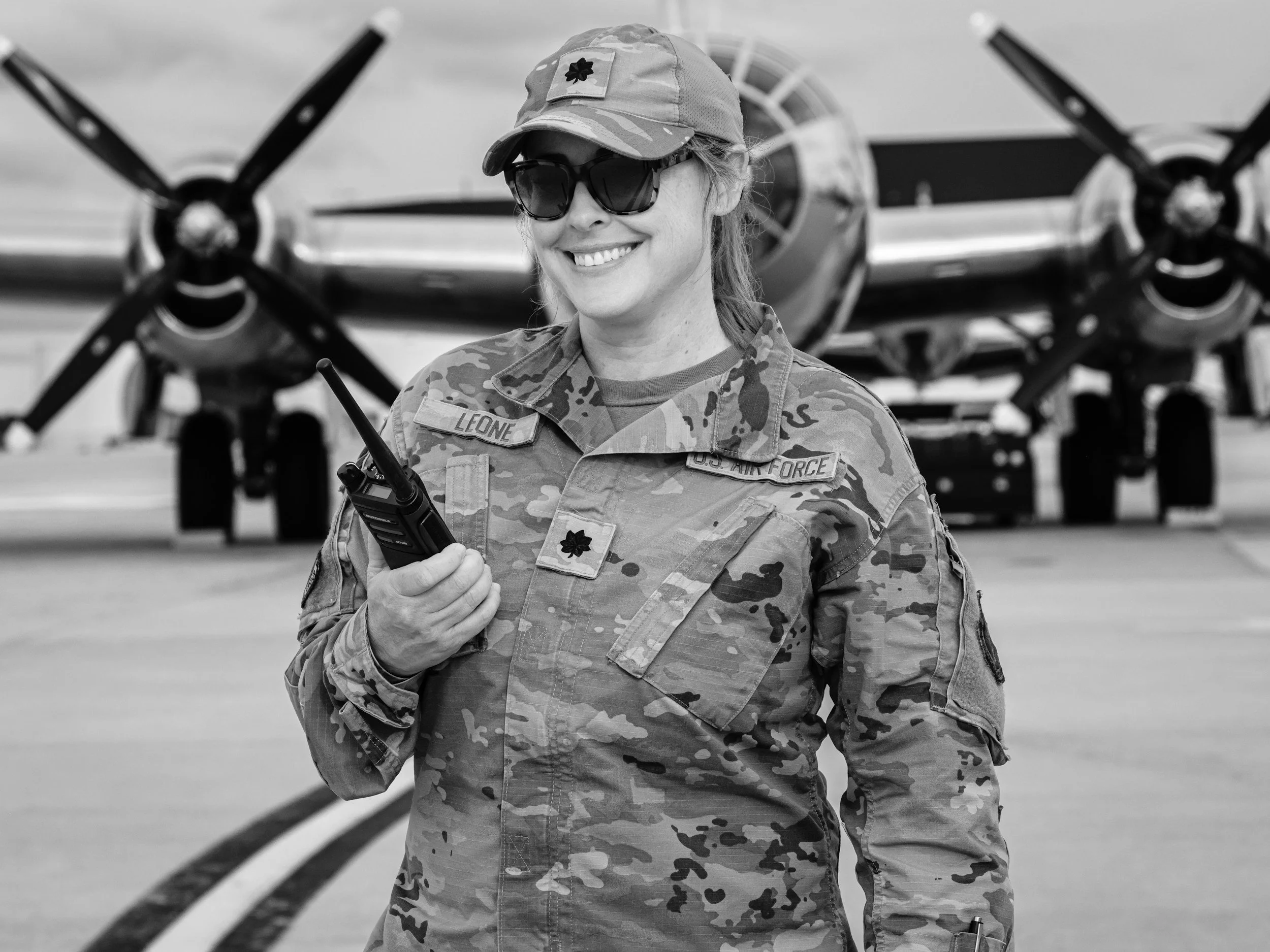 A female military personnel in camouflage uniform and sunglasses holding a walkie-talkie, standing in front of a propeller airplane.