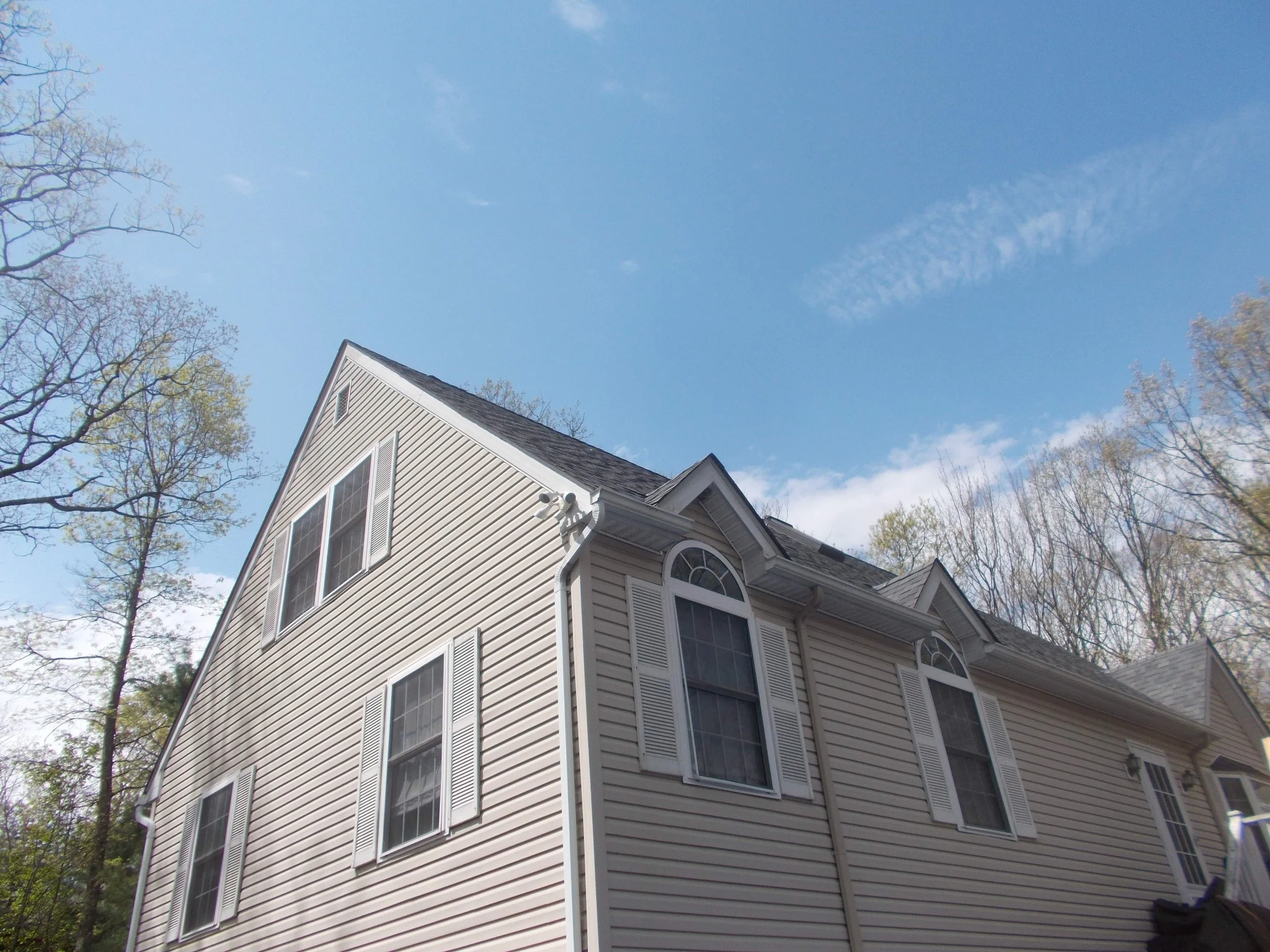 A two-story house in Lords Valley PA with beige vinyl siding and white window shutters, surrounded by trees, under a partly cloudy blue sky.