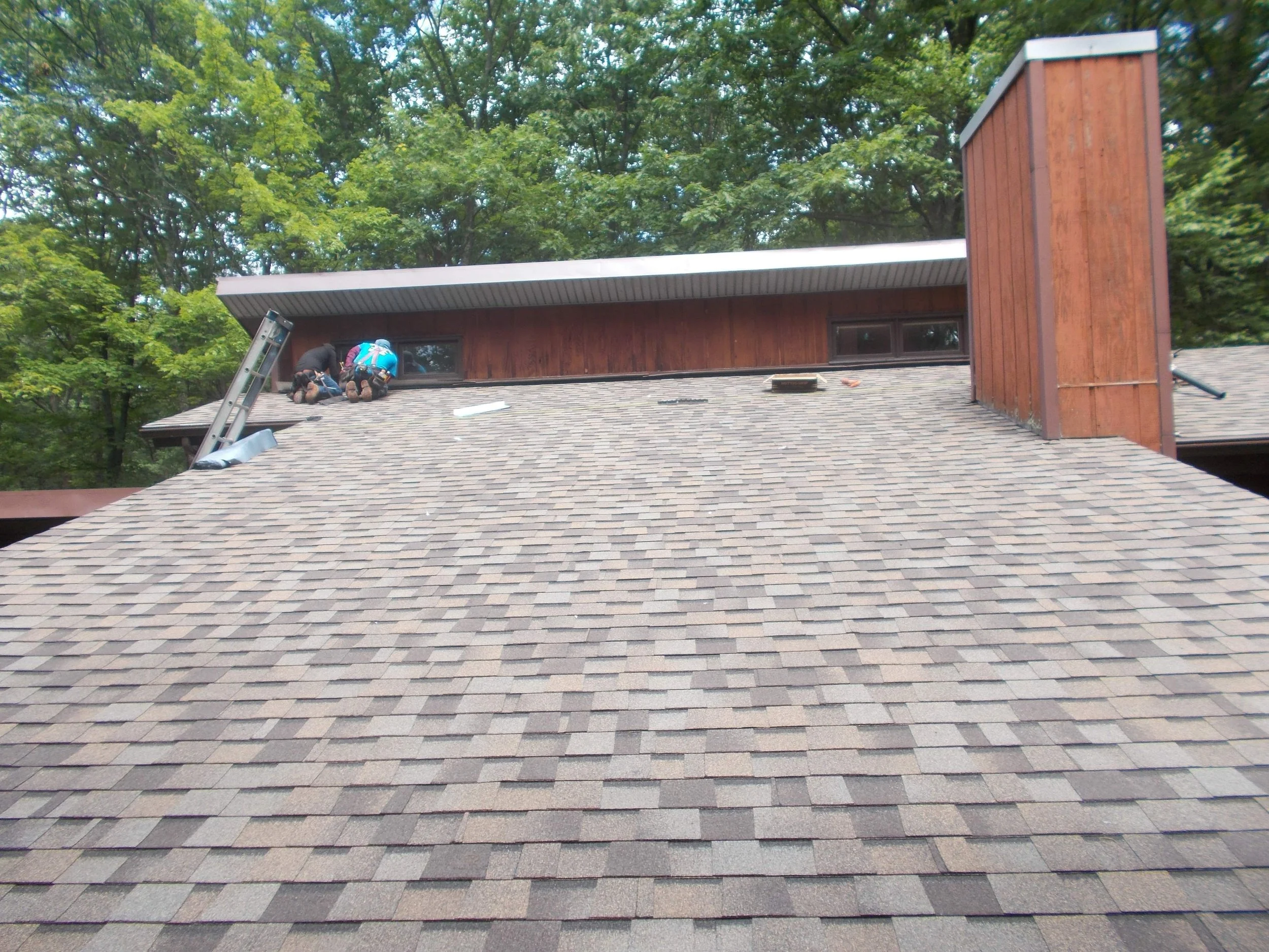 Two roofers working on the roof of a house that has brown shingle roofing and a wooden exterior, in Lords Valley PA.