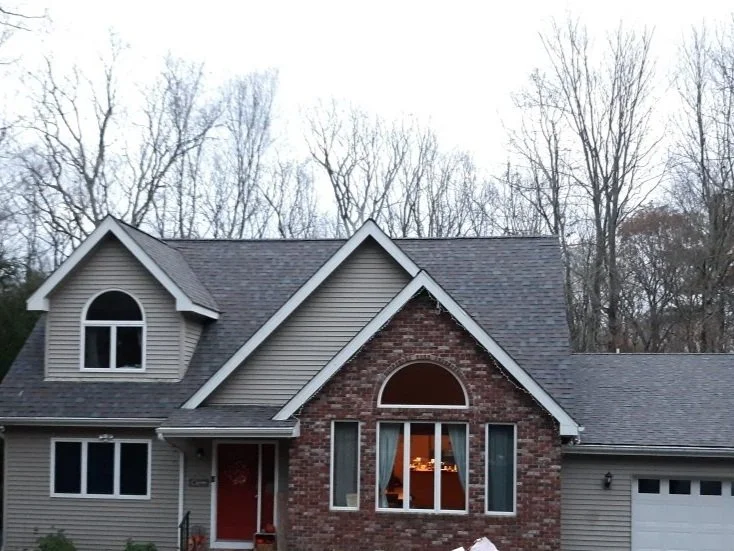 Front view of a two-story house in Milford PA with beige siding, a brick facade, and a gabled roof, surrounded by bare trees, with a red front door and a large front window.