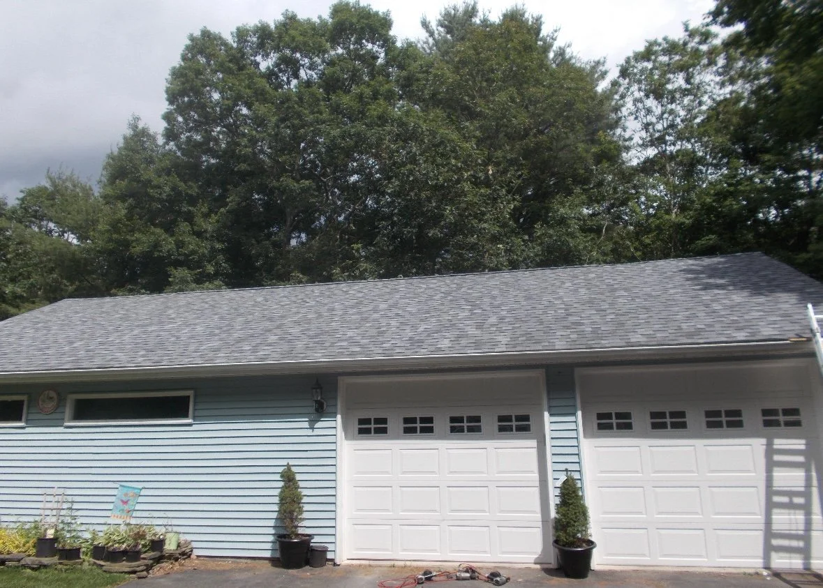 A house in Bushkill PA with gray shingled roof and blue siding, featuring a double garage with white doors and small windows at the top. 