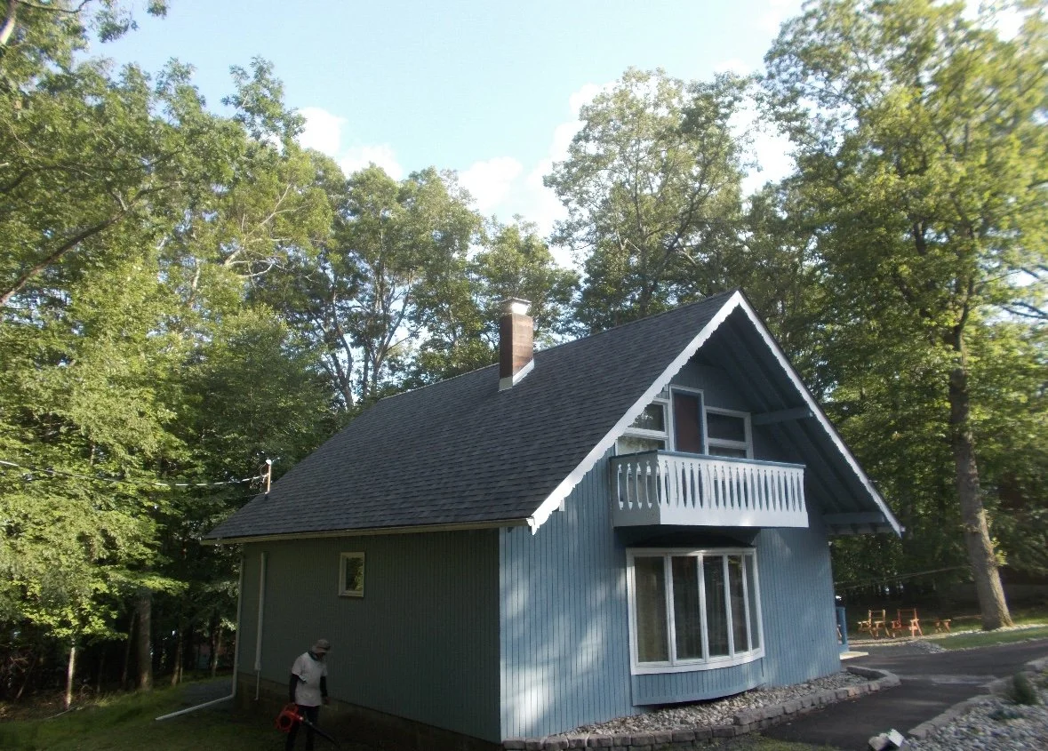 A two-story blue house in Milford PA, Gold Key, with a gabled roof and a small front porch, surrounded by trees under a bright sky.