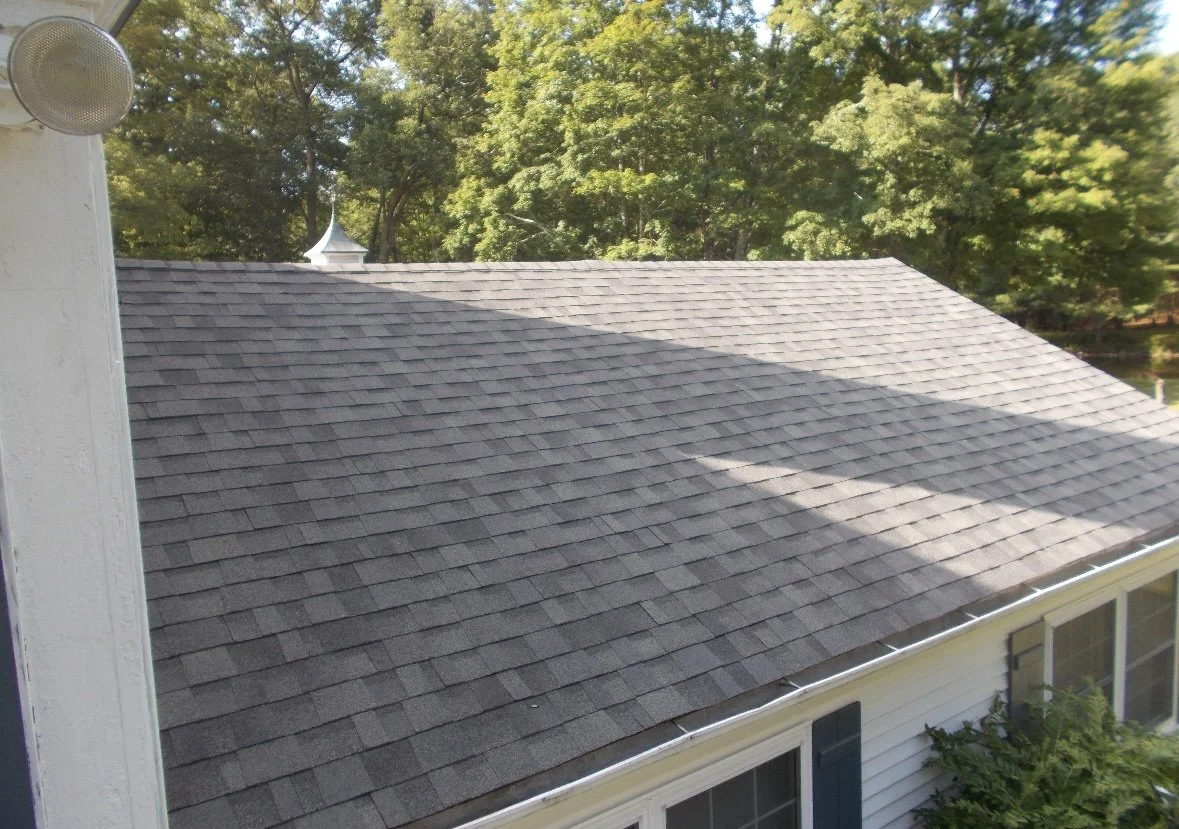 View of a house roof in Milford PA with gray shingles, part of the house's white exterior, and surrounding green trees.