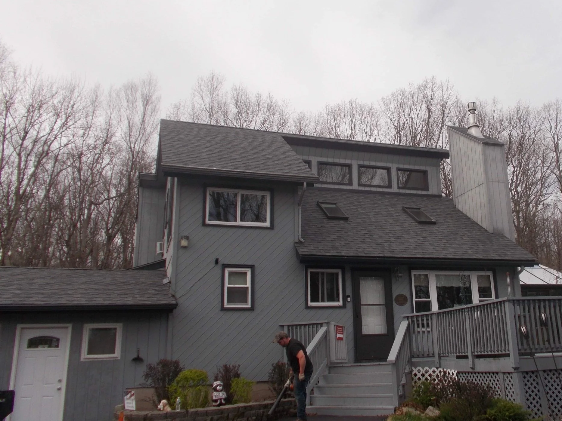 A new roof in Milford PA. Multi-story gray house with a wood deck, surrounded by leafless trees under an overcast sky.