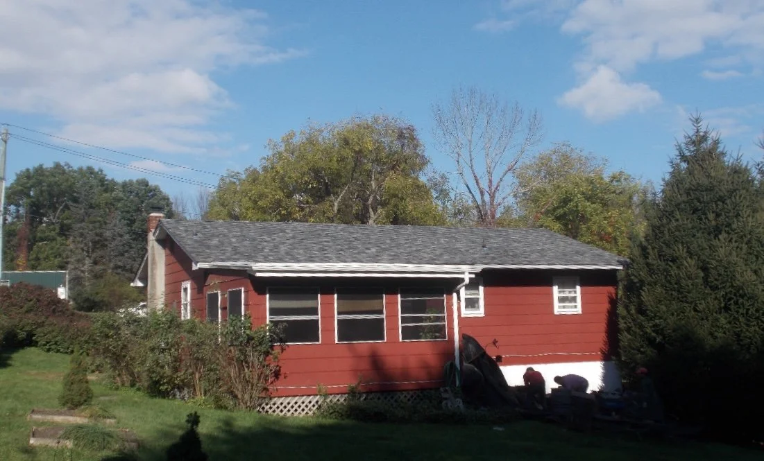 Red house in Montague NJ with screened porch, gray roof, surrounded by trees and bushes, under a blue sky with clouds.