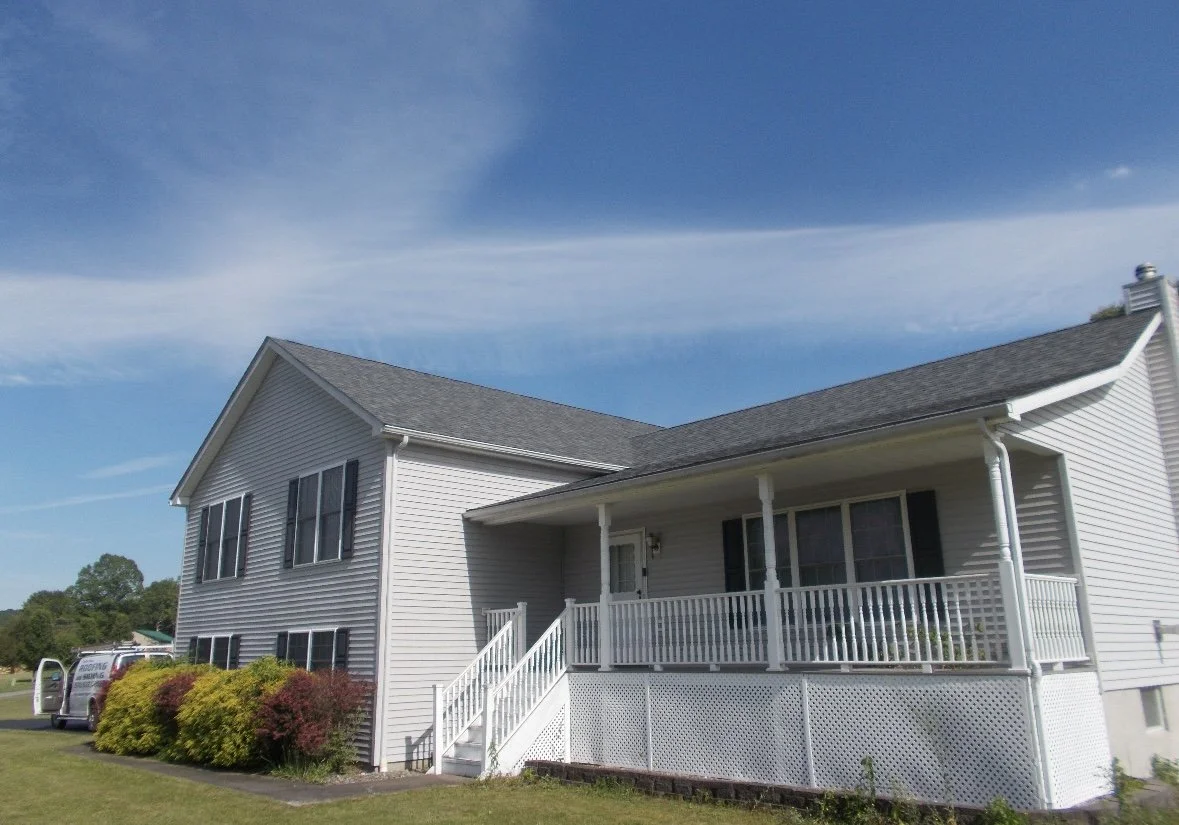 A two-story house in Hawley PA with white siding, black shutters, and a covered front porch with a white railing. There are bushes and a lawn in front, and a white van parked nearby. The sky is partly cloudy with a blue background.