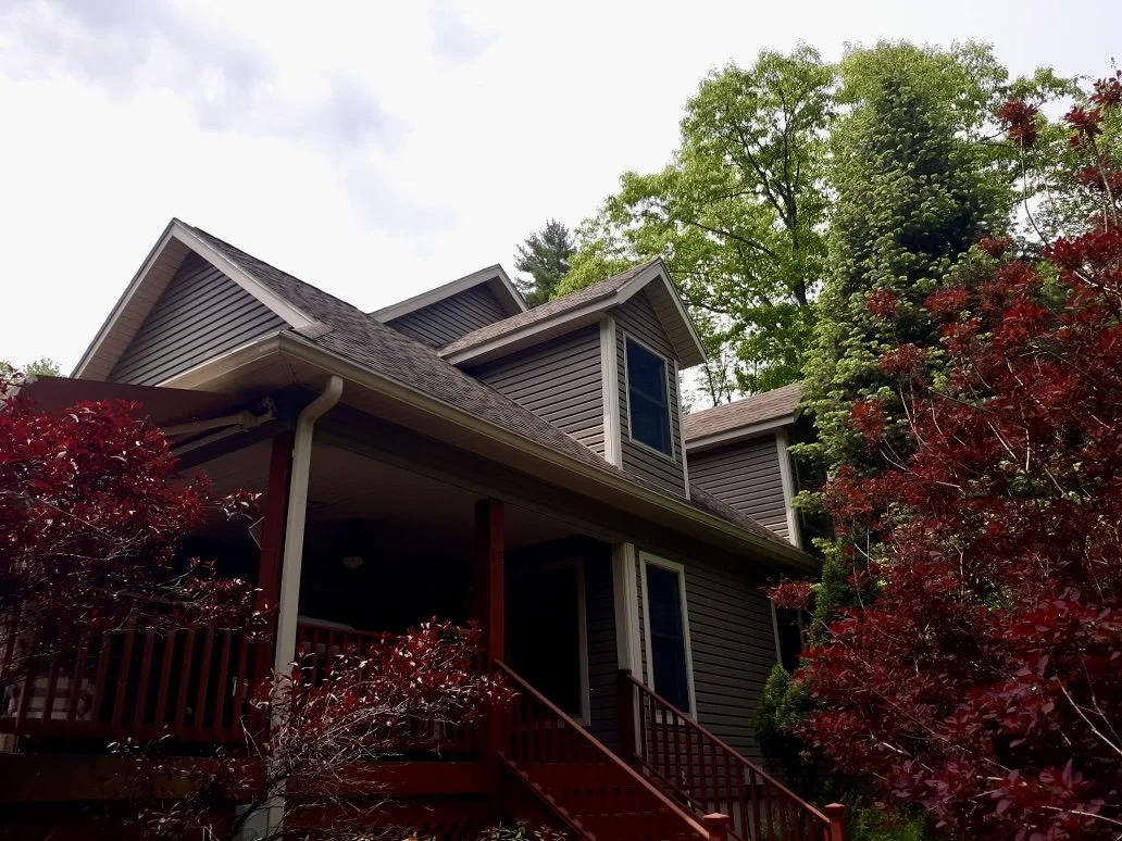 A two-story house in Milford PA with dark gray siding, a brown shingled roof, and a porch with red stairs surrounded by trees and red shrubbery.