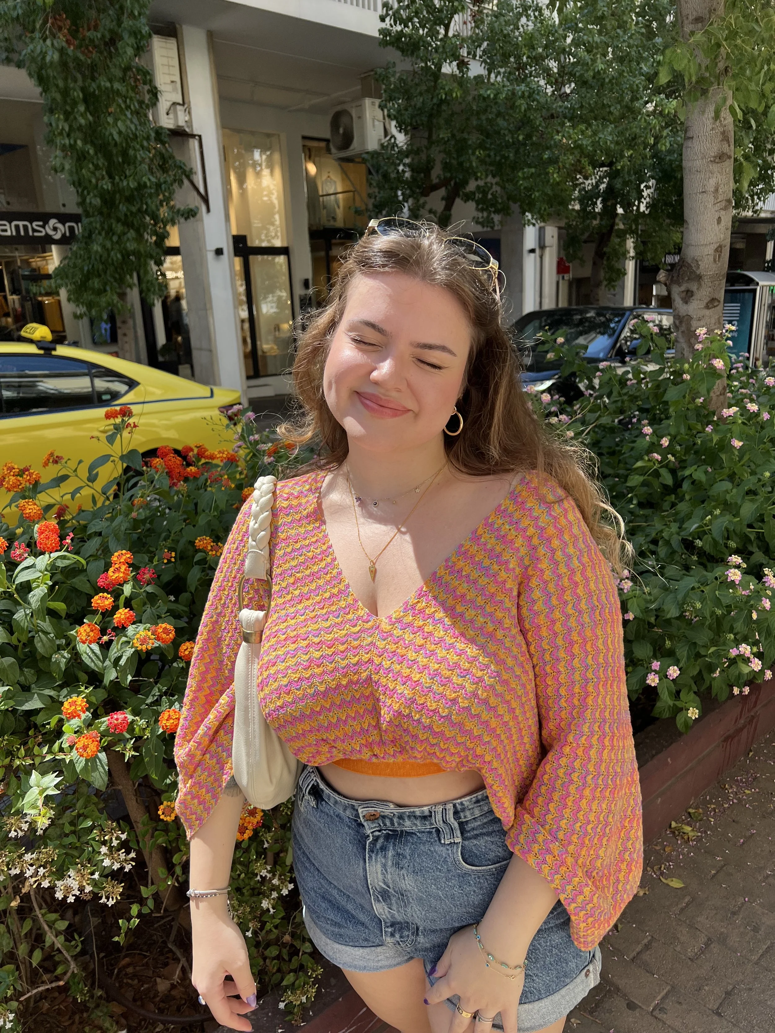 A young woman standing on a sidewalk with closed eyes, smiling, wearing a colorful pink and orange zigzag patterned top, high-waisted denim shorts, sunglasses on her head, and jewelry, surrounded by blooming flowers and trees.