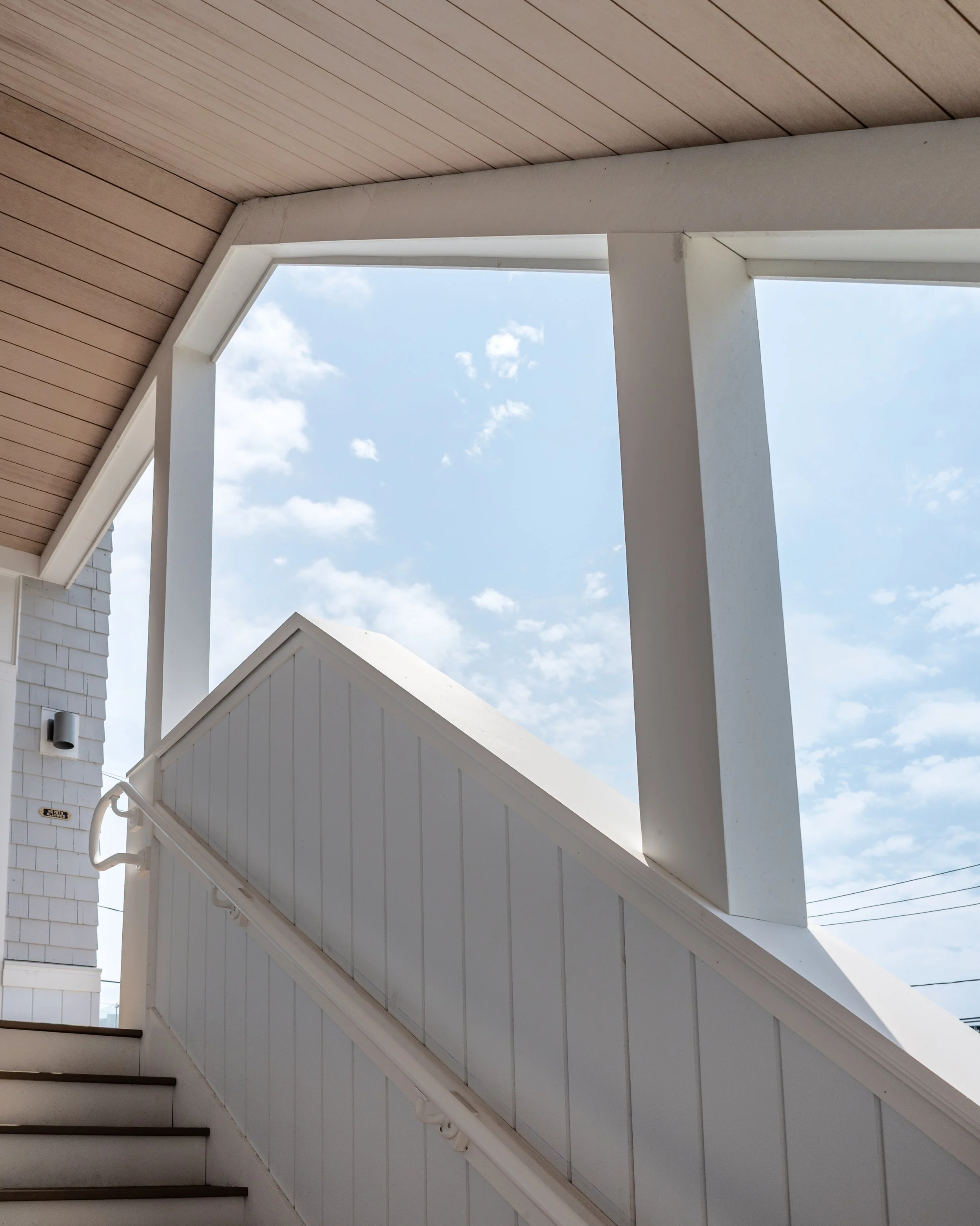 Part of a staircase with black steps, adjacent to a white wall with a metal pipe. Above, a white triangular frame surrounds an open sky with a few scattered clouds.