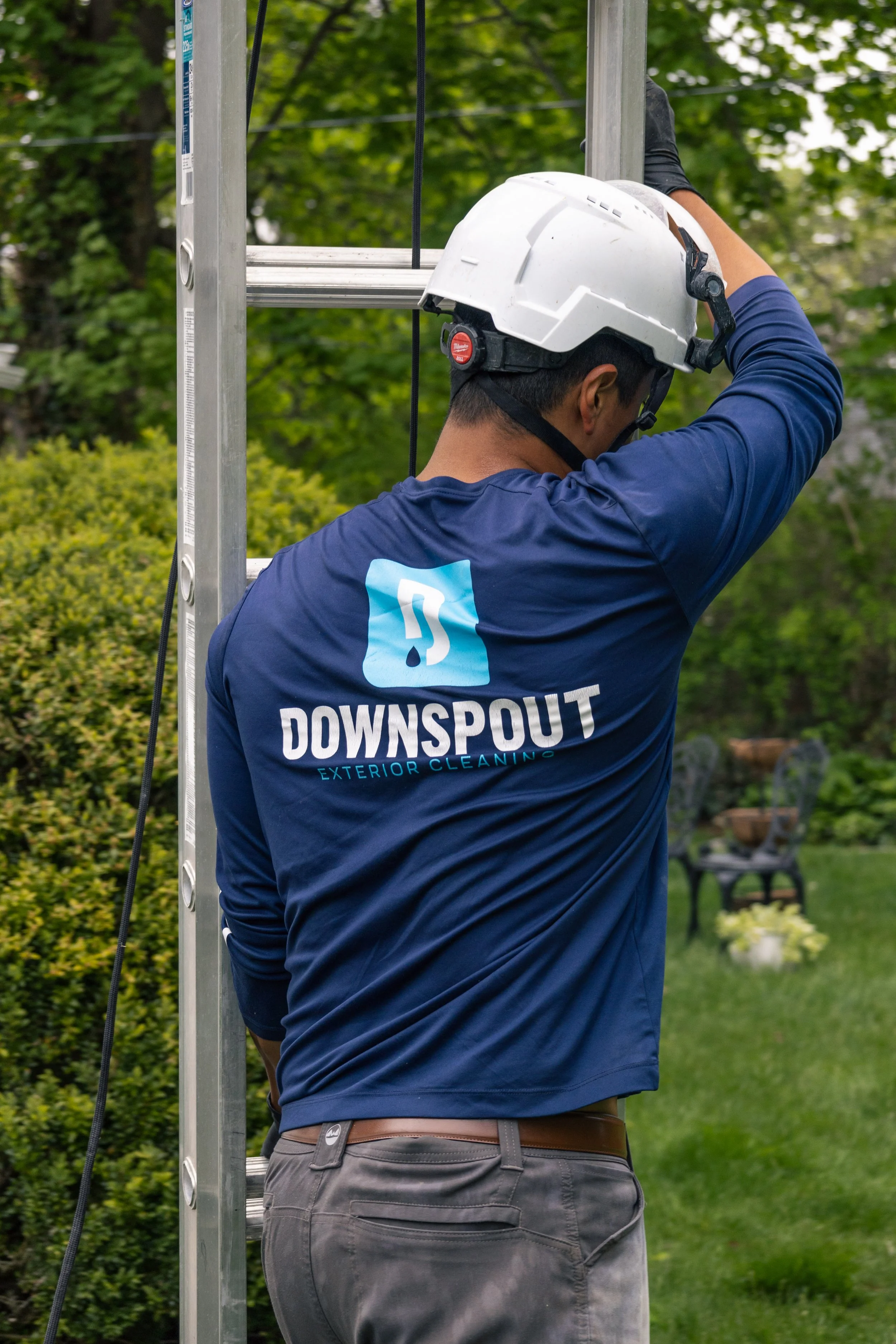 A person wearing a white safety helmet and a blue shirt with the "Downspout Exterior Cleaning" logo is working on a metal ladder outdoors, with green trees and a garden in the background.