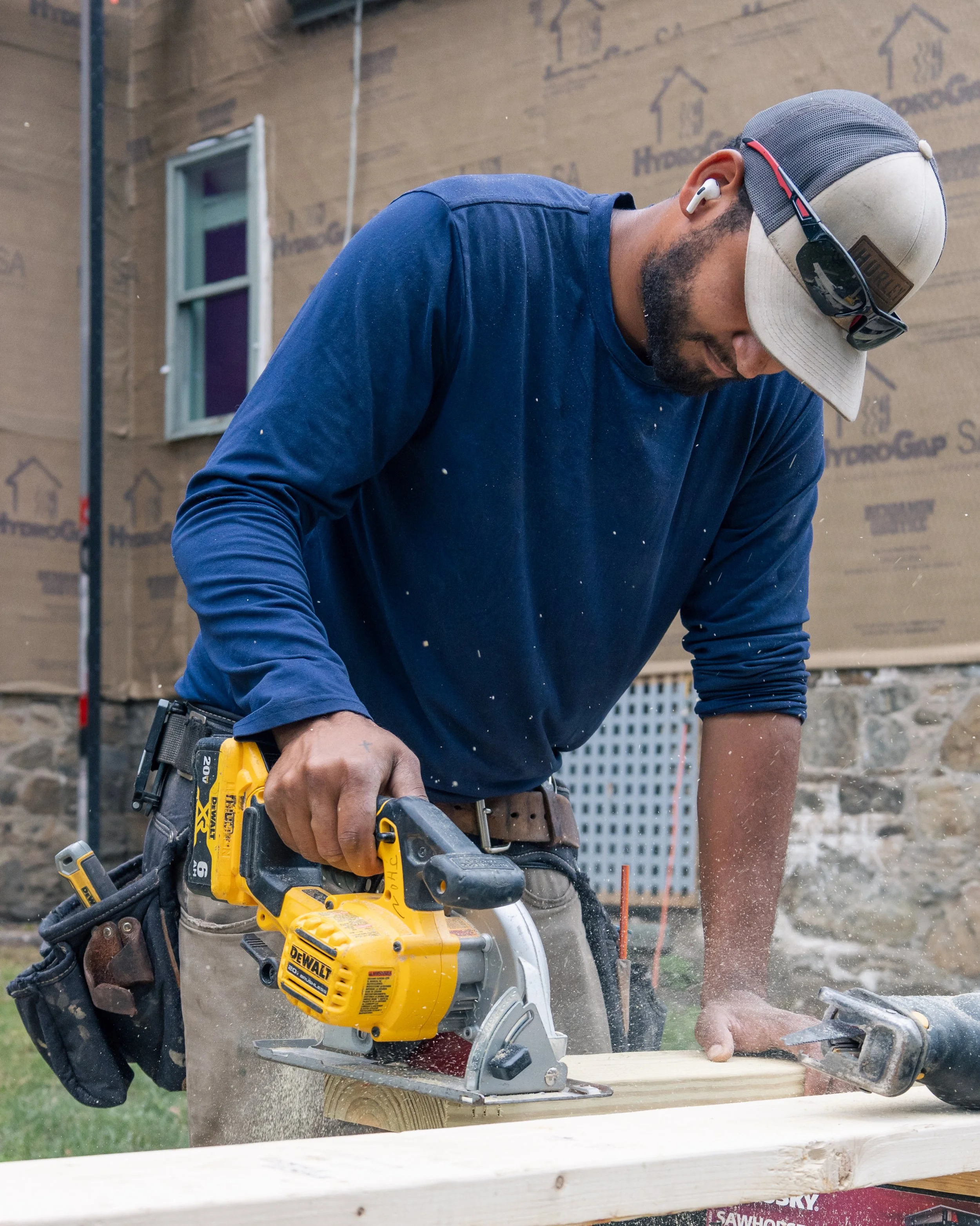 A man using a yellow DeWalt circular saw to cut wood outdoors, with a background of a building under construction.