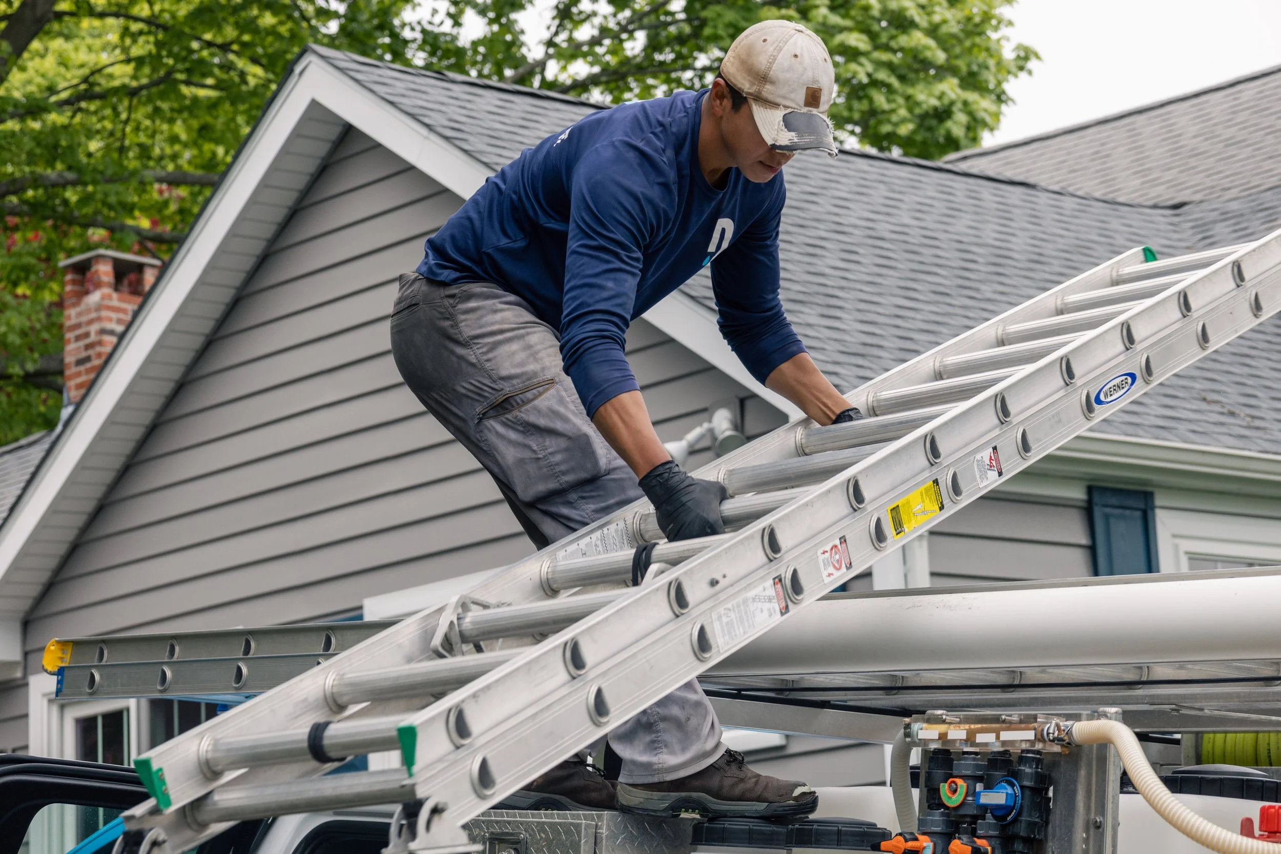 A worker wearing a beige cap and a blue shirt working on an outdoor roof with a ladder, near neighboring houses and green trees.