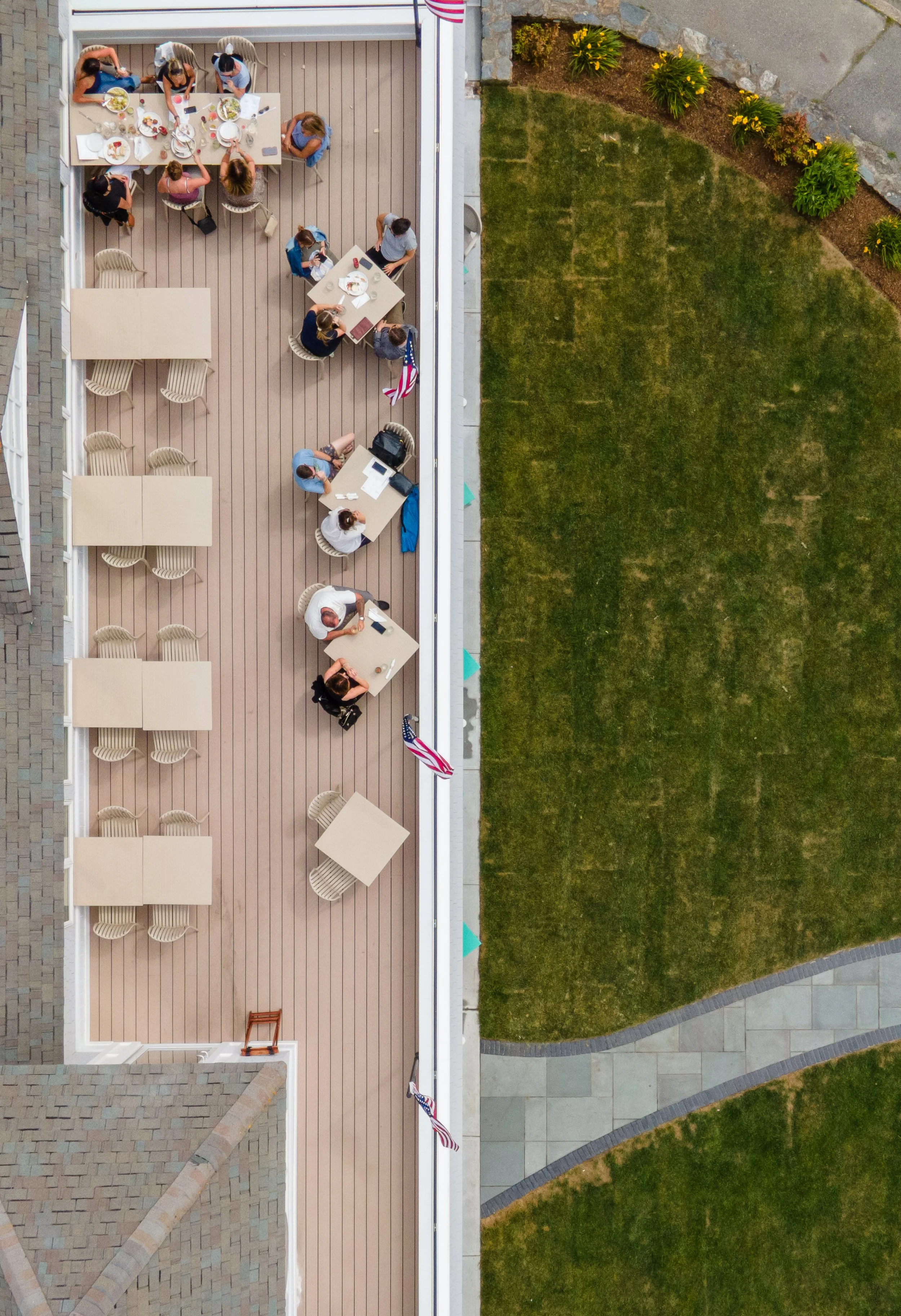 An aerial view of a rooftop deck with several tables and chairs, and a group of people sitting and dining. The deck has a wooden floor, and the surrounding area includes a grassy lawn with garden beds and patio stones.