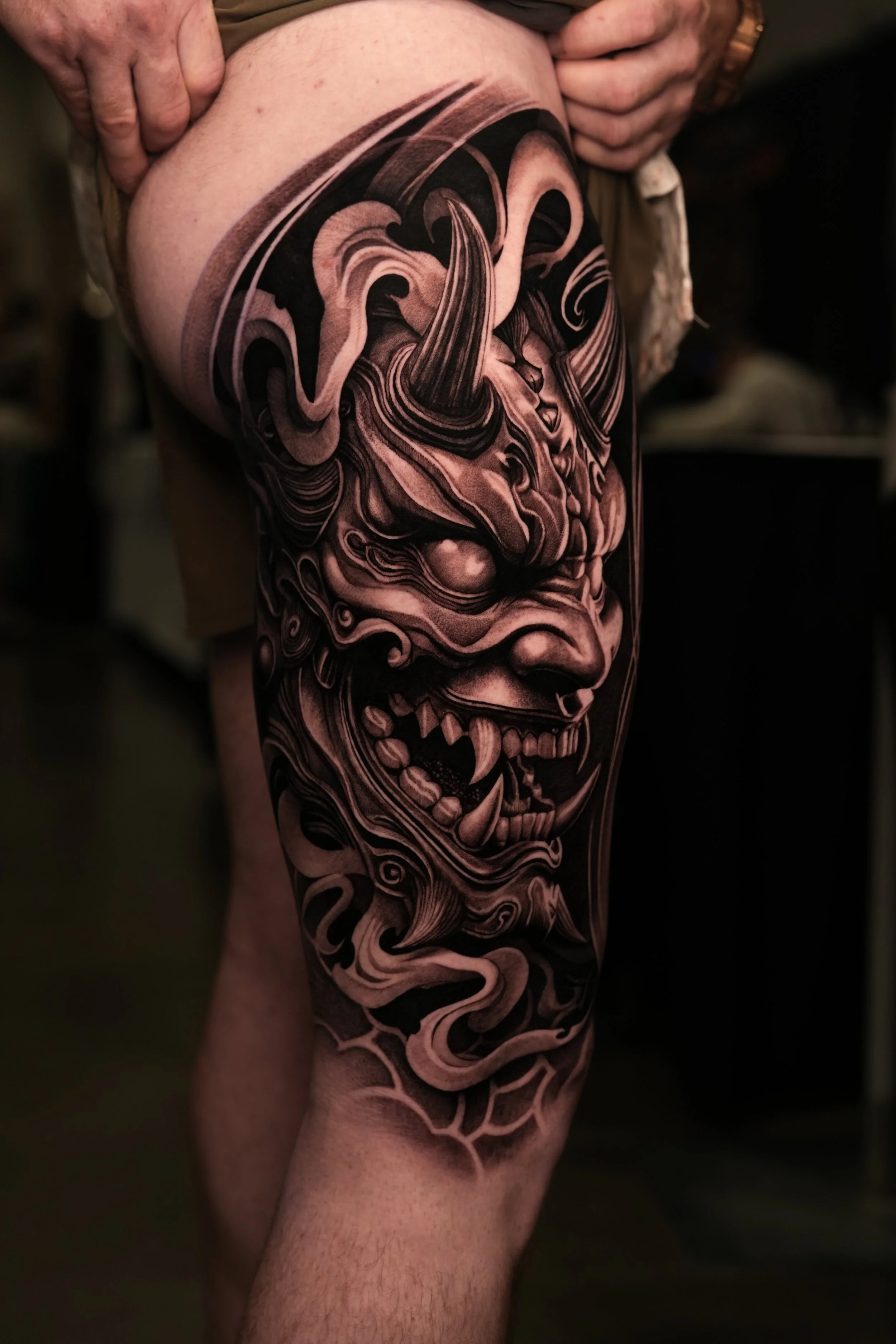 Close-up of a detailed black and gray tattoo of a fierce demon face with horns and sharp teeth on a person's thigh.
