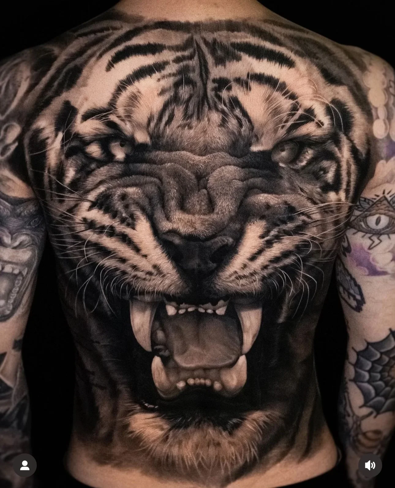 Realistic tiger face tattoo covering a person's back, with an open mouth and detailed stripes. Surrounding tattoos appear on the arms.