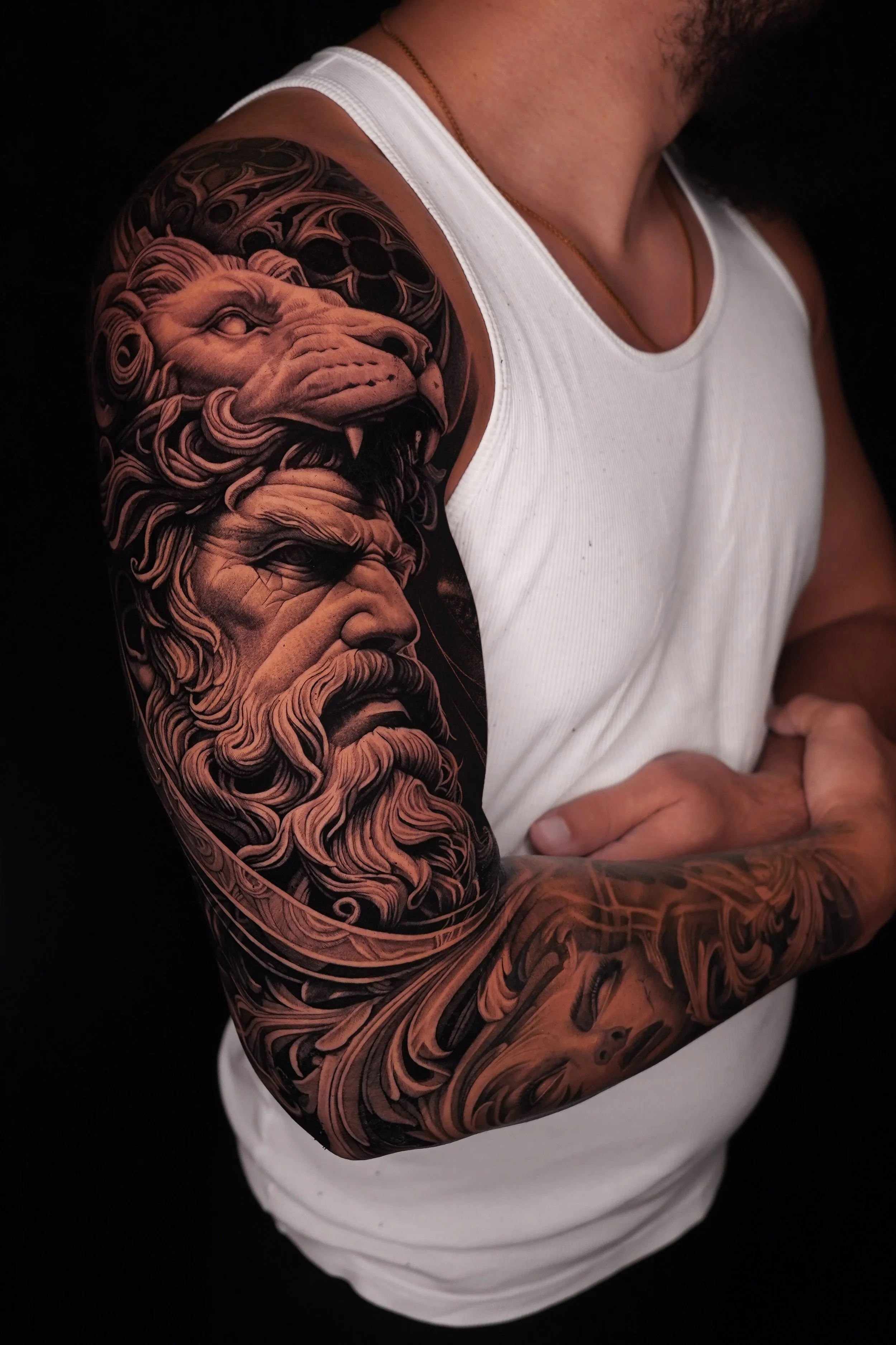 A person's arm with a detailed black and gray tattoo of a mythological figure with a beard, eyes closed, and intricate patterns, along with a lion's head with a mane and fierce expression, on the upper arm and shoulder.