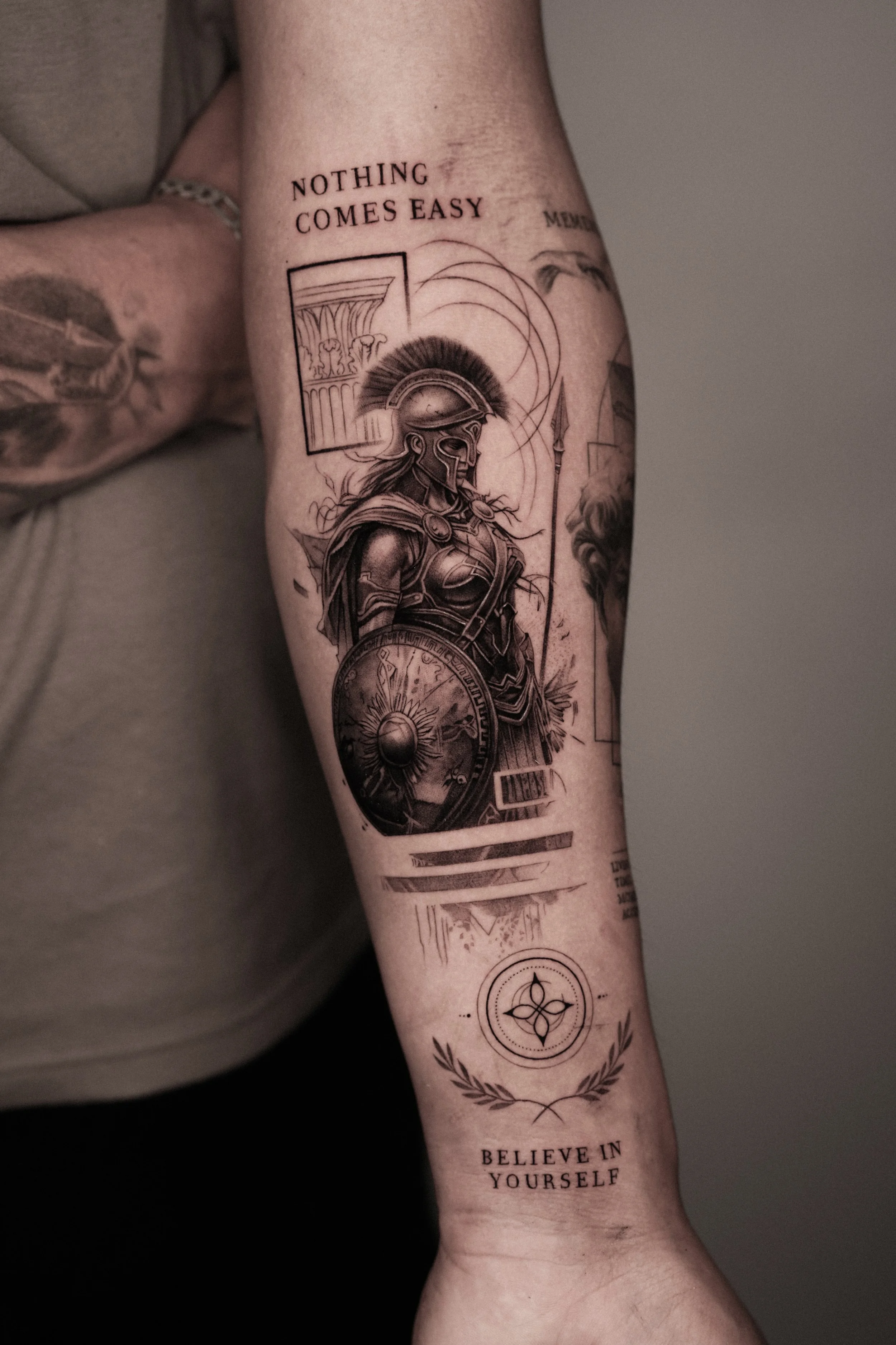 Black and gray tattoo of a female warrior in ancient armor holding a shield and spear on a person's arm, with inspirational quotes and symbols surrounding her.