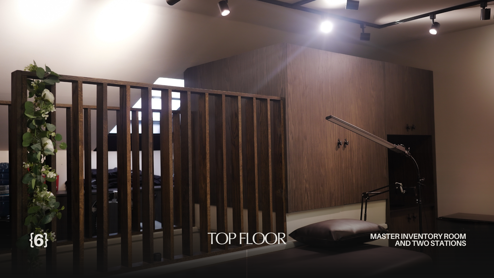 The image shows a modern interior space with wooden elements. There is a wooden railing with green and white decorative plant on the left. The ceiling has black track lights. The wall in the background is made of wood paneling, with a small dark open