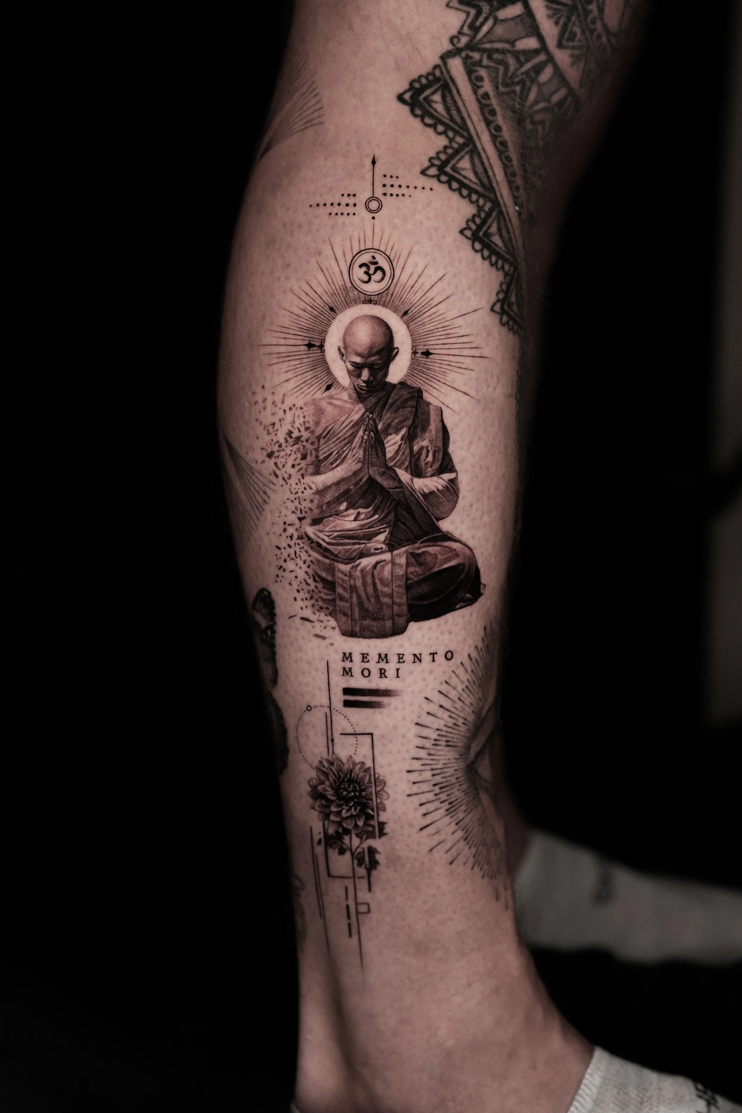 Tattoo of a praying Buddhist monk with a halo, surrounded by radiating lines, a circle with an 'om' symbol, geometric designs, a flower, and the Latin phrase 'Memento Mori'.