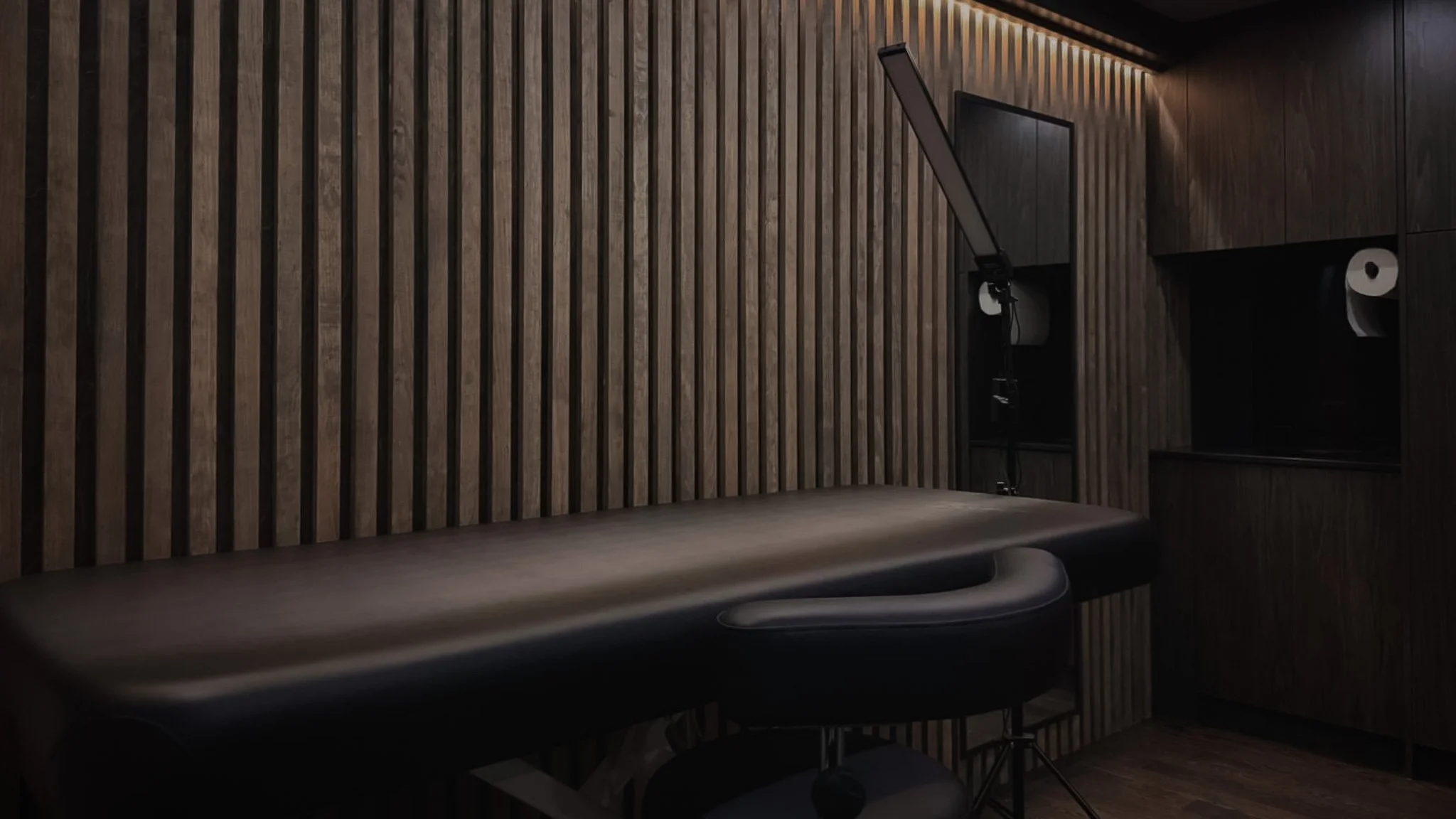 Modern tattoo studio with a wooden slatted wall, black massage chair, and overhead lighting.