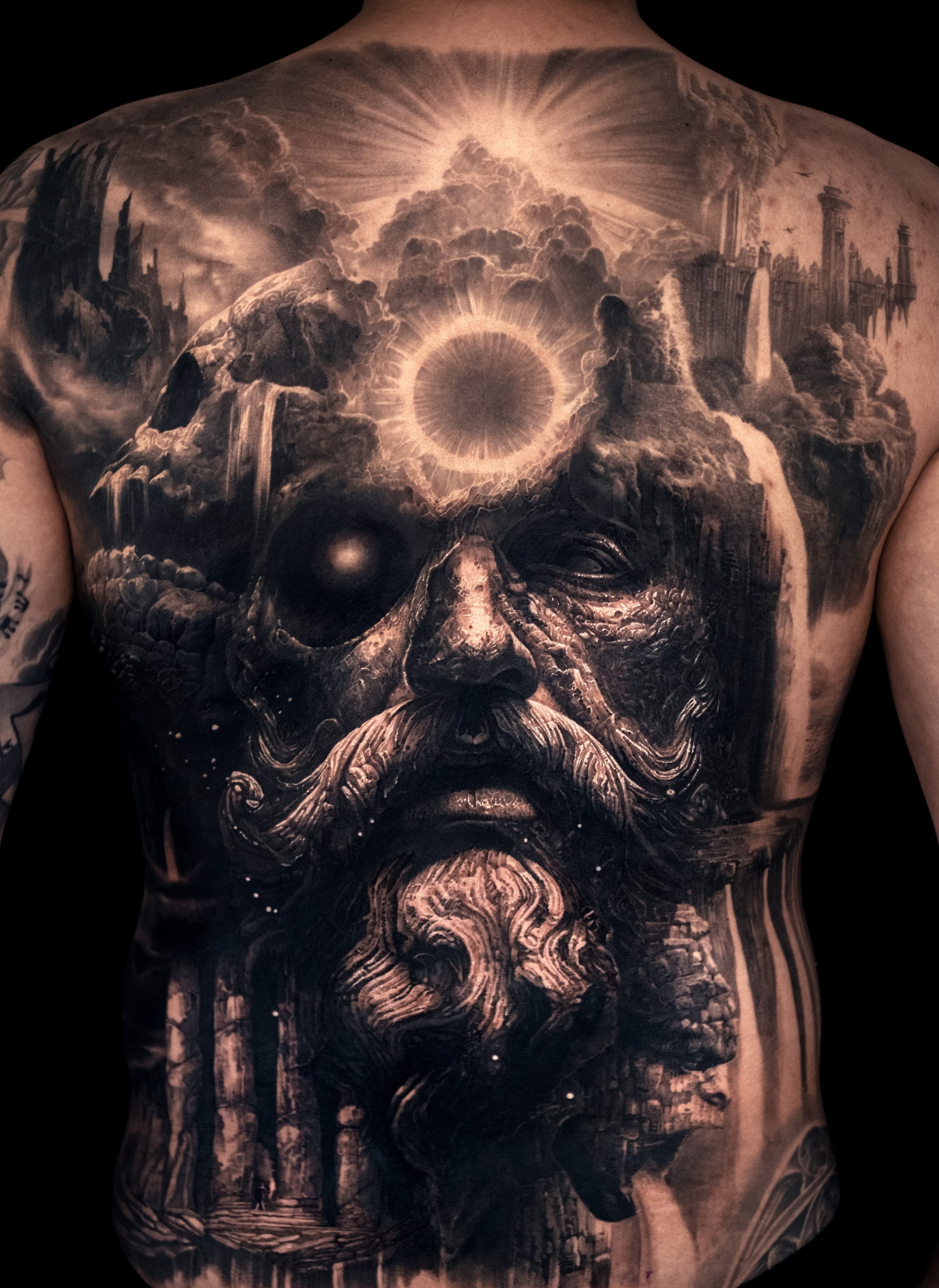 Black and gray tattoo of a surreal face on a person's back, featuring a bearded figure blended with landscapes and celestial elements like clouds and a dark sun.
