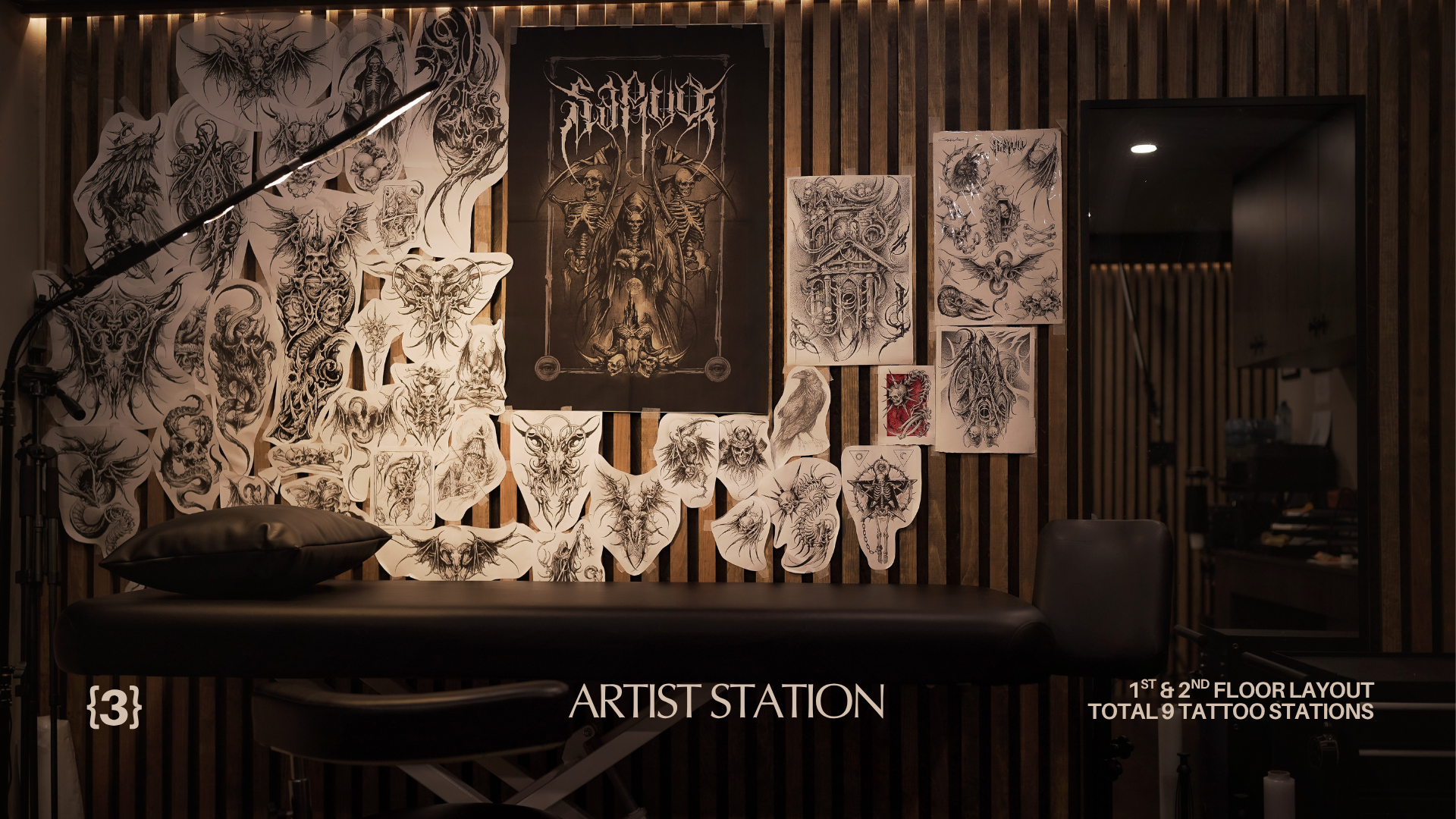 Tattoo artist's station with various sketches and artwork on the wall, including detailed black and white designs of mythical and fantasy creatures. The station has a black massage table, a pillow, and a black chair, with wooden paneling in the backg
