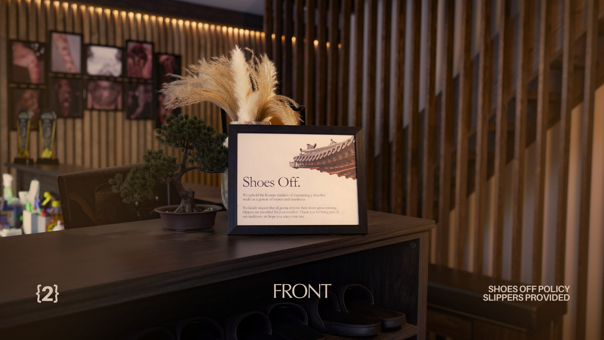 A front desk area with a framed sign that says "Shoes Off" and explains the store's policy on removing shoes upon entering. The desk has a small bonsai tree and decorative pampas grass in the background, with a wooden wall and stairs behind.