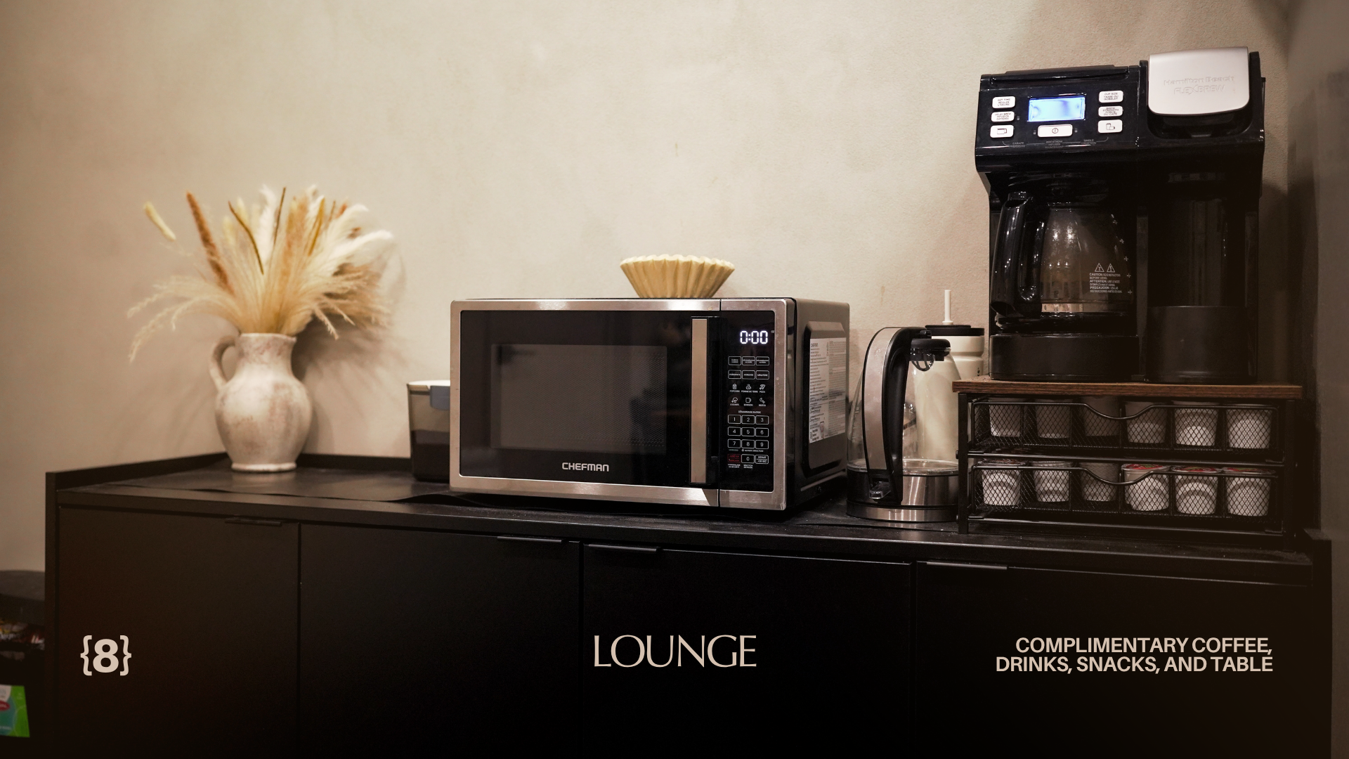 Coffee station with a vase of dried flowers, microwave, coffee maker, and a container on a black table with 'LOUNGE' written on it, in a room offering complimentary coffee, drinks, snacks, and table.