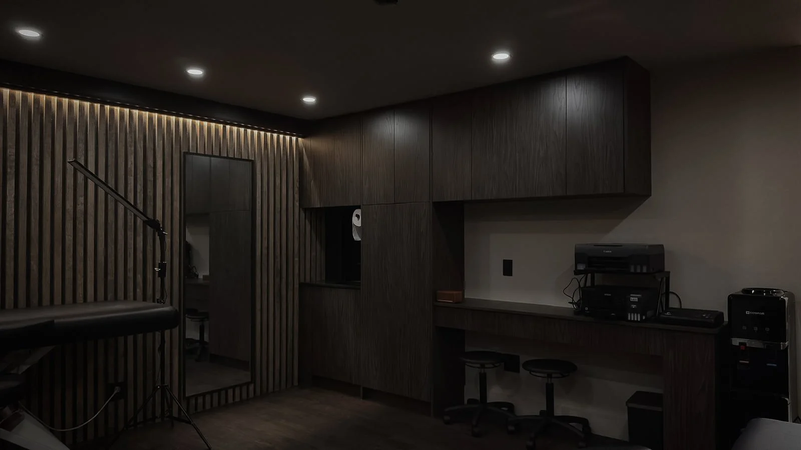 Dimly lit office space with dark wood paneling, a large mirror, ceiling lights, a printer, a water dispenser, and a treatment chair.