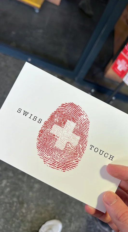 A hand holding a white card with a red fingerprint and a white Swiss cross in the center. The card has the words 'SWISS TOUCH' printed around the fingerprint.