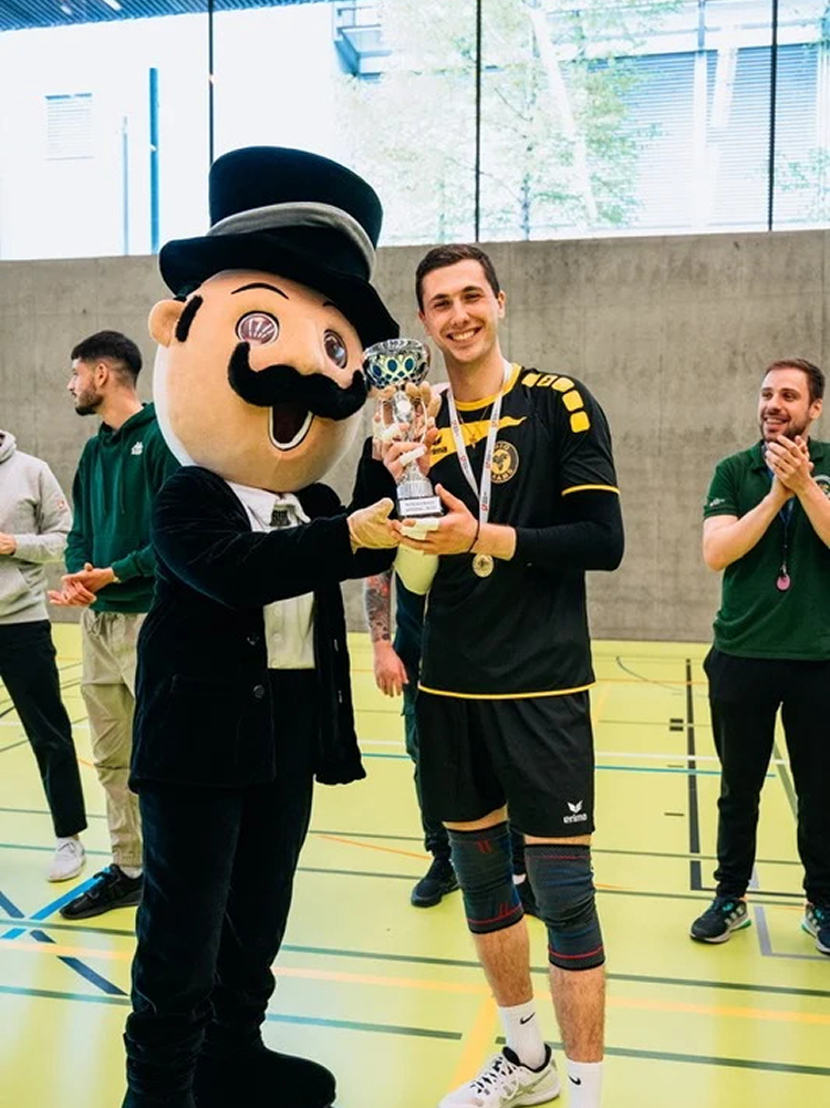 A young man in a black and yellow sports uniform holding a trophy, standing next to a mascot in a black suit, large top hat, and mustache, inside a gymnasium with other people clapping in the background.