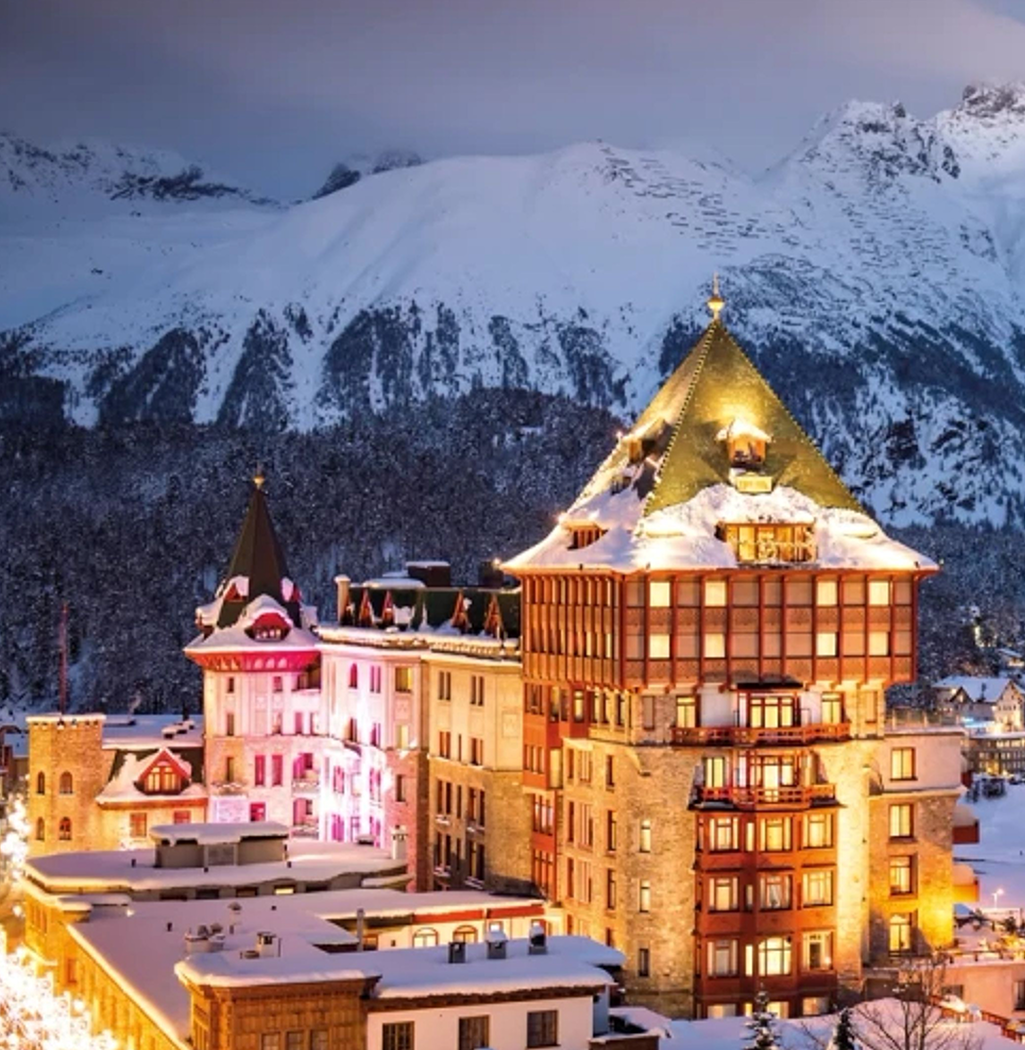 Snow-covered castle with pointed roofs and warm lights, set against a backdrop of snowy mountains at dusk.