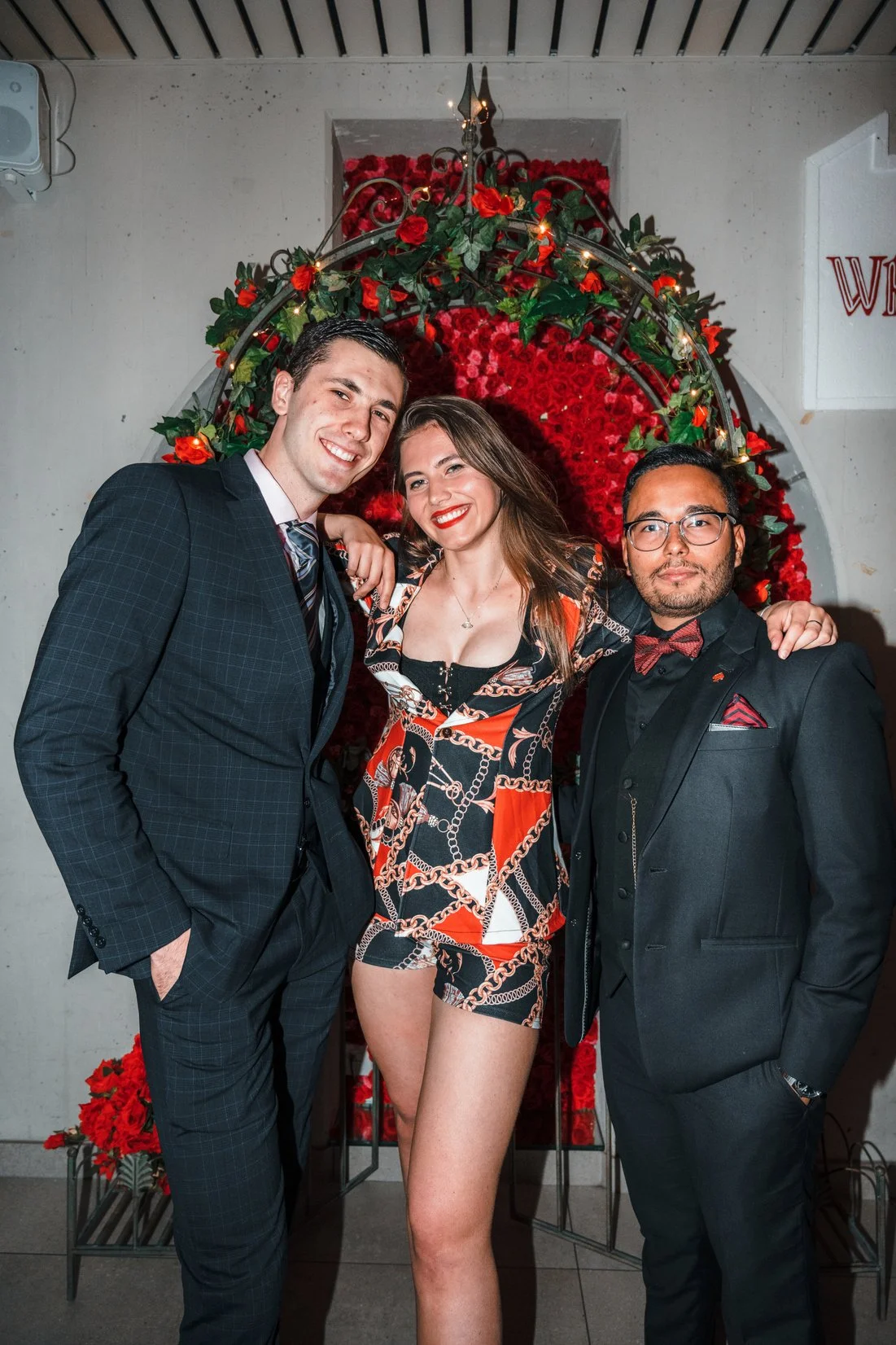 Three people dressed in formal attire standing in front of a floral backdrop with red roses and greenery, smiling for the camera.