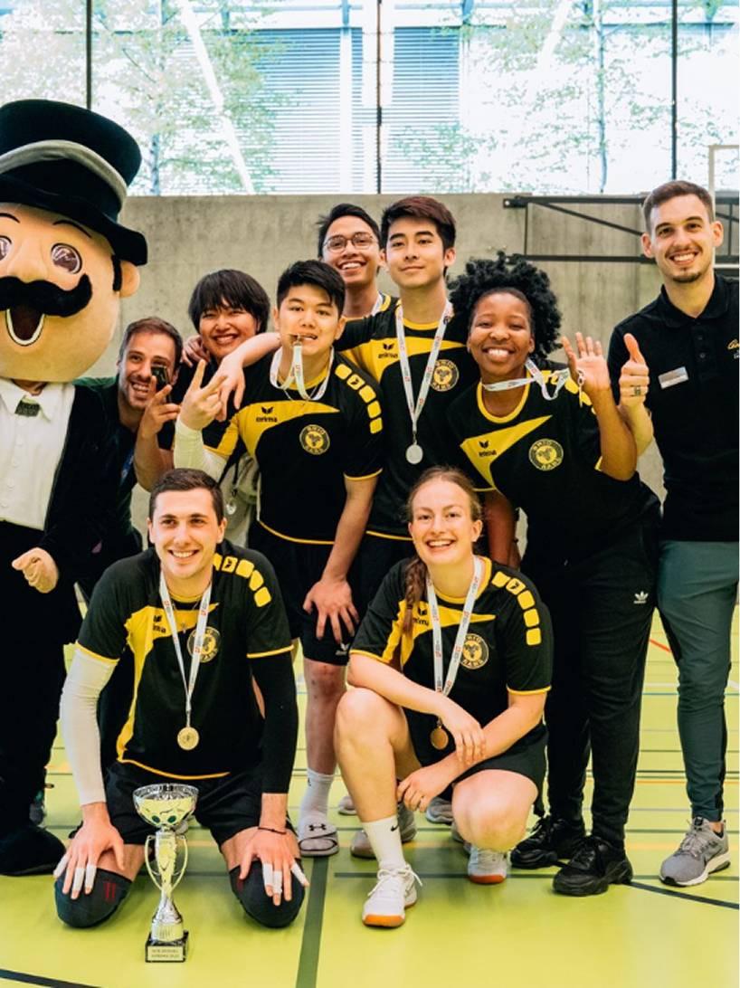 Group of young athletes in black and yellow soccer uniforms with medals, celebrating with a large mascot and a trophy in an indoor sports facility.
