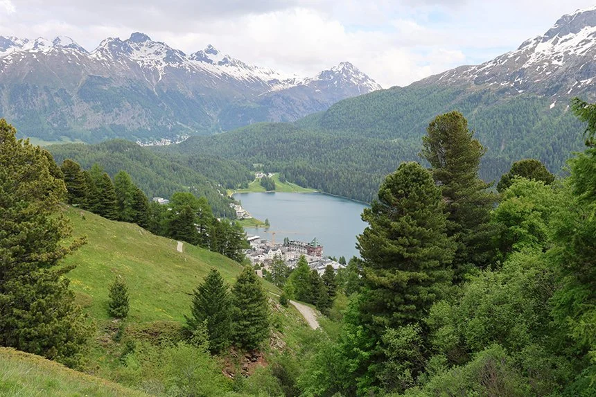 A scenic view of a lake surrounded by green forests and mountains, with a small town or village visible near the water and snow-capped peaks in the distance.