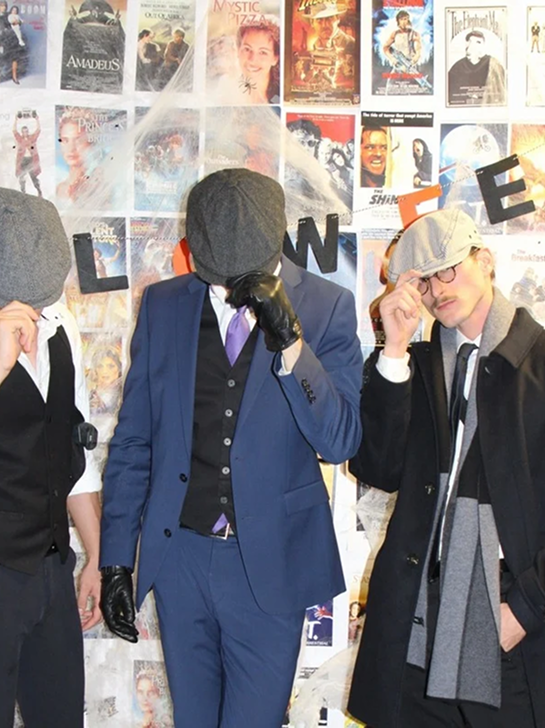 Three men in vintage and formal clothing posing in front of a collage of movie posters and the word 'LOVE' spelled out on black and orange letters.