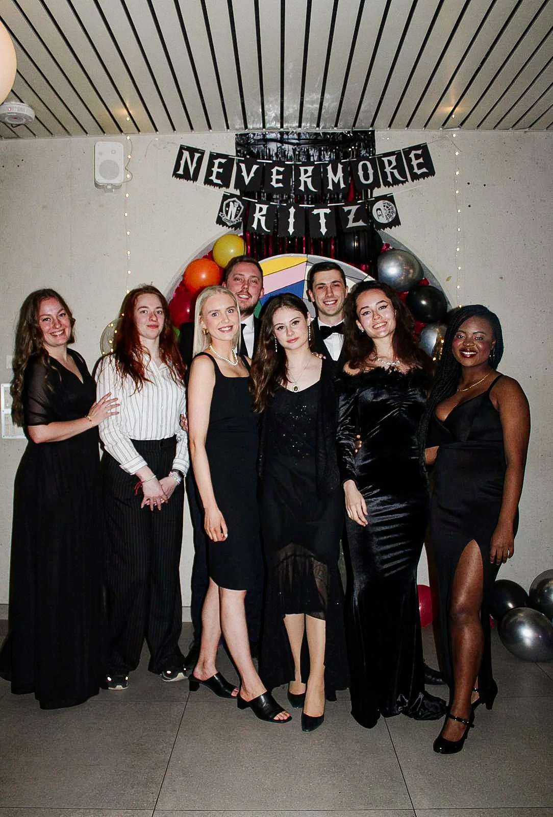 Group of eight young adults dressed in formal black attire, standing together and smiling at a celebration, with colorful balloons and a banner that reads 'NEVERMORE' and 'RITZ' in the background.