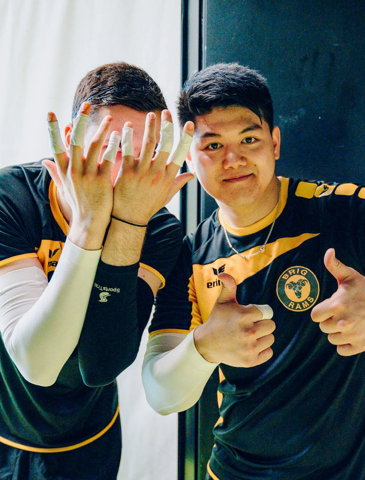 Two young men in black and yellow sports jerseys taking a selfie. One is covering his face with his hands covered in fingerless gloves, and the other is giving a thumbs-up and pointing at his friend.