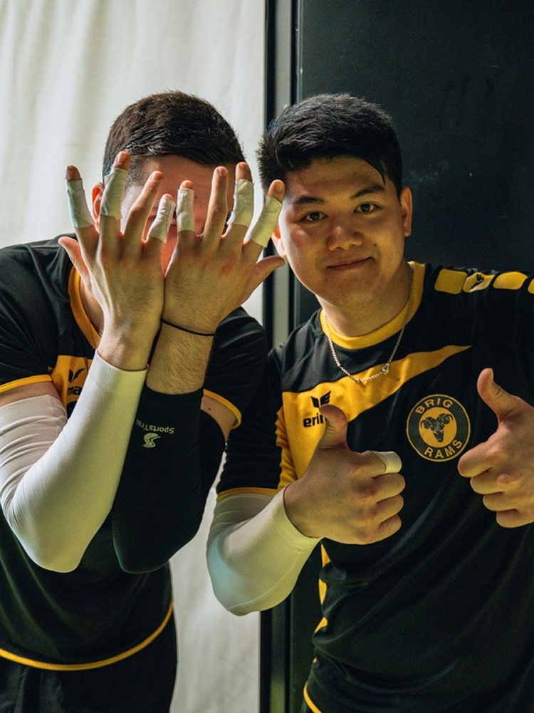 Two young men in sports jerseys with a team logo, one with bandaged fingers and the other giving thumbs up, inside a room with a curtain and wall.