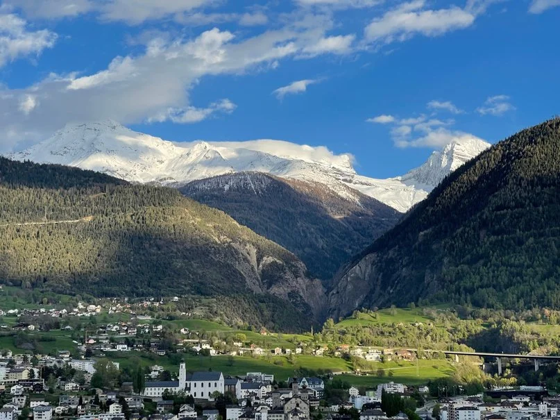 A scenic view of snow-capped mountains over a green valley with a small town and a bridge.