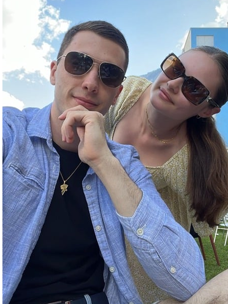 A young man and woman outdoors, both wearing sunglasses. The man is wearing a blue shirt and a necklace with a cross, and is resting his chin on his hand. The woman is wearing a yellow patterned top and is leaning in close to the man. The background shows a partly cloudy sky and a blue and white building.