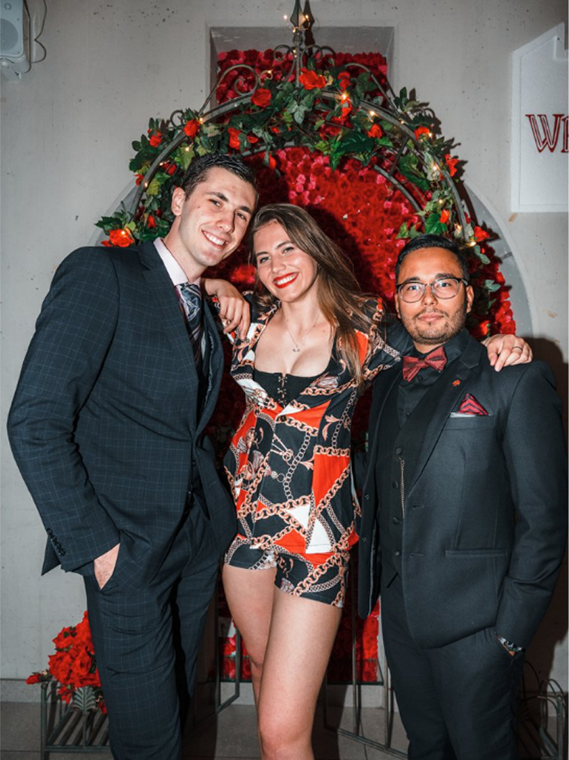 Three people dressed in formal attire standing in front of a floral heart-shaped backdrop decorated with red roses. They are smiling and posing for a photo at a festive event.